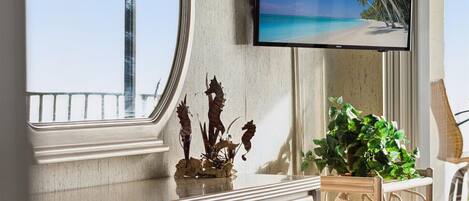Una habitación con un ventanal grande, un televisor de pantalla plana mostrando una escena de playa, una planta decorativa y un mueble de madera.