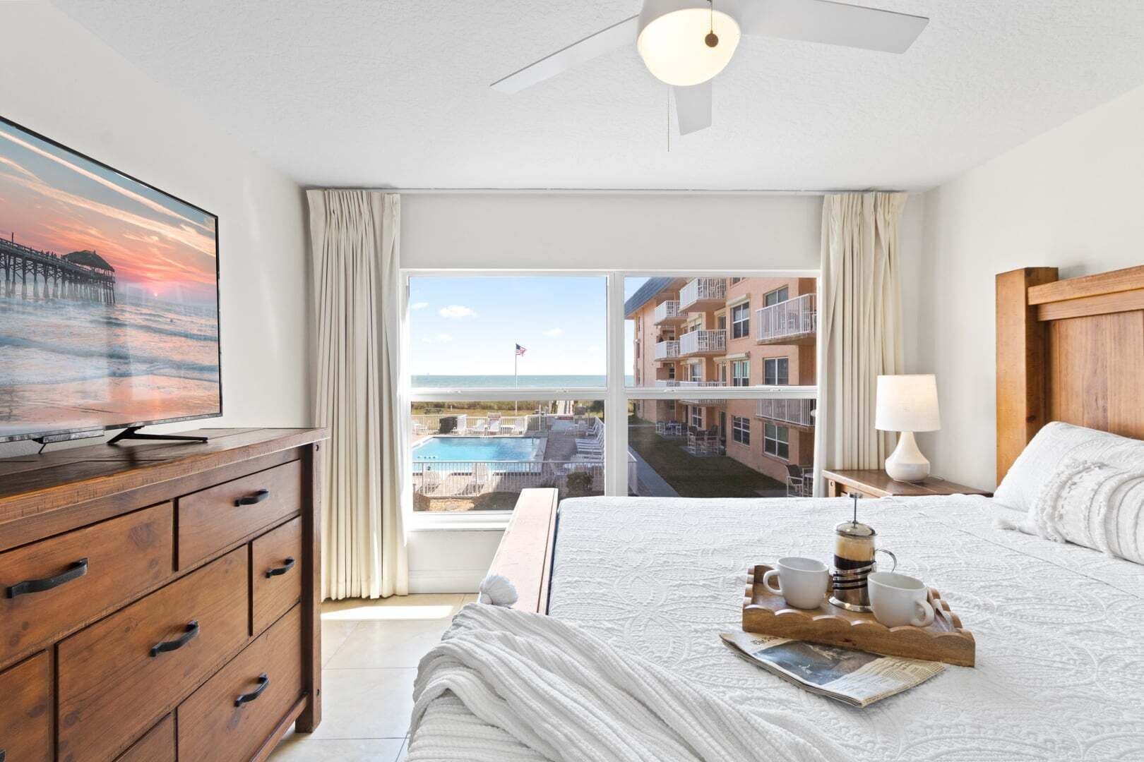 A hotel room with a large bed, a wooden dresser, a television, a window with a view of a building and a pool, and a ceiling fan.