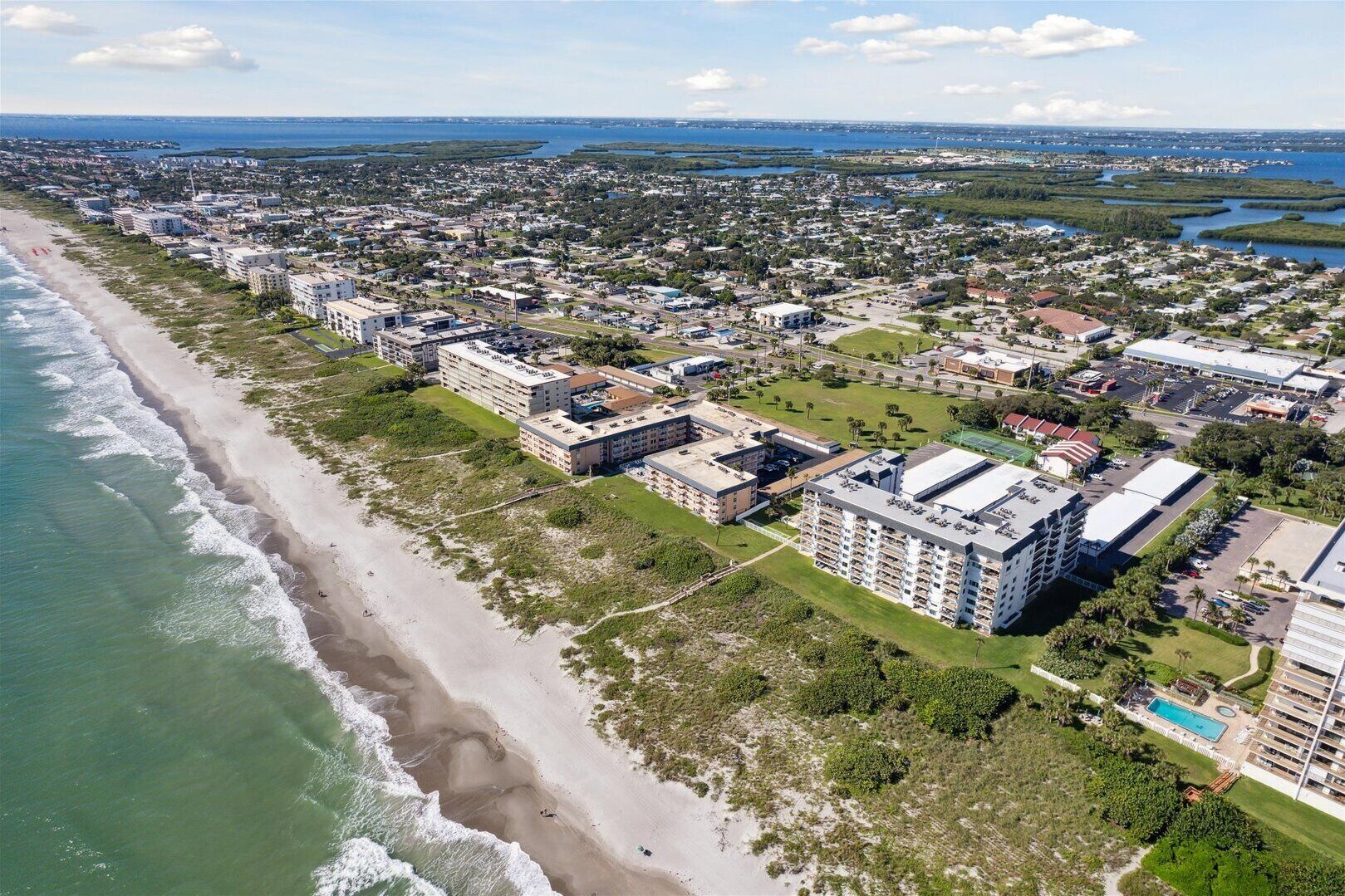 An aerial view of a coastal area with a beachfront hotel, multiple buildings, and a marina.