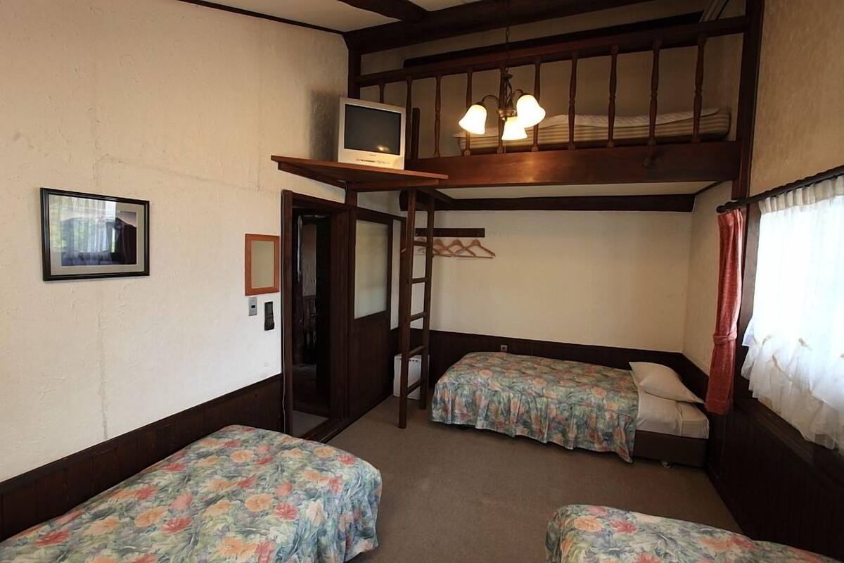 Habitación tipo dormitorio con dos camas individuales, un televisor en un estante, una escalera y una ventana con cortinas.