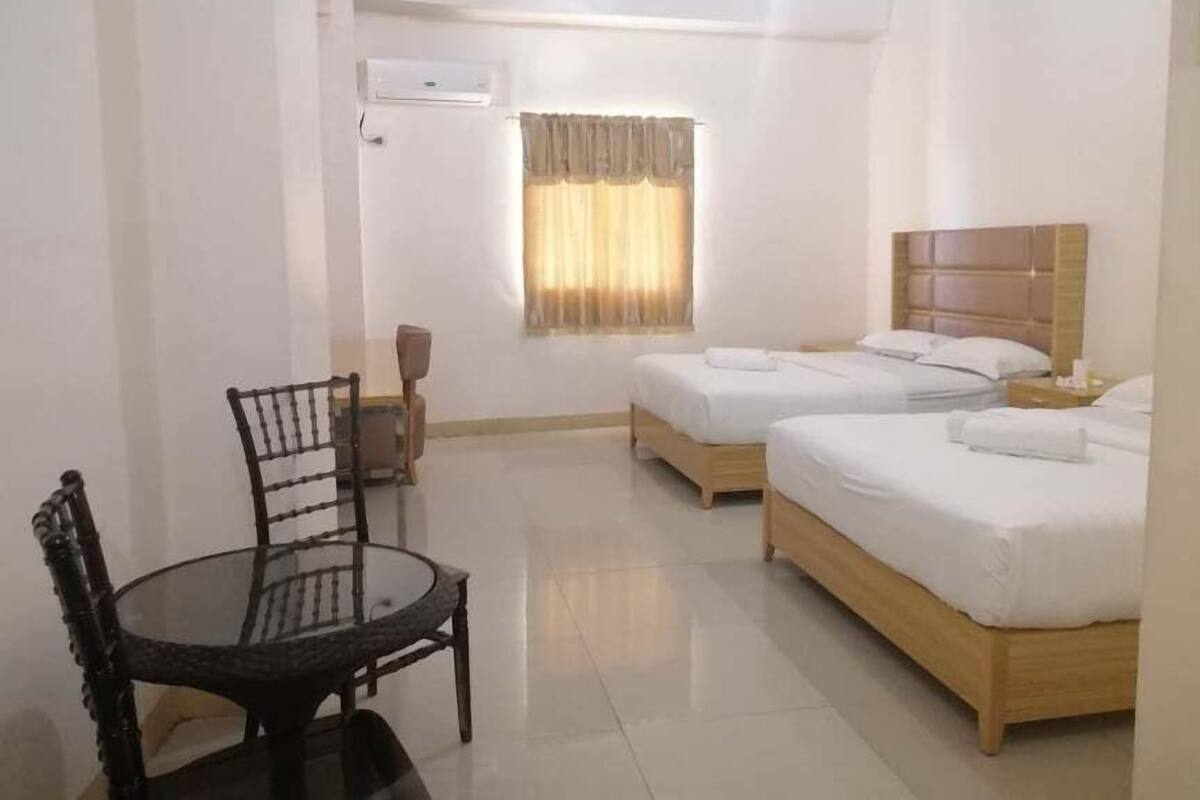 A hotel room with two beds, a small table with a chair, and a window with curtains.