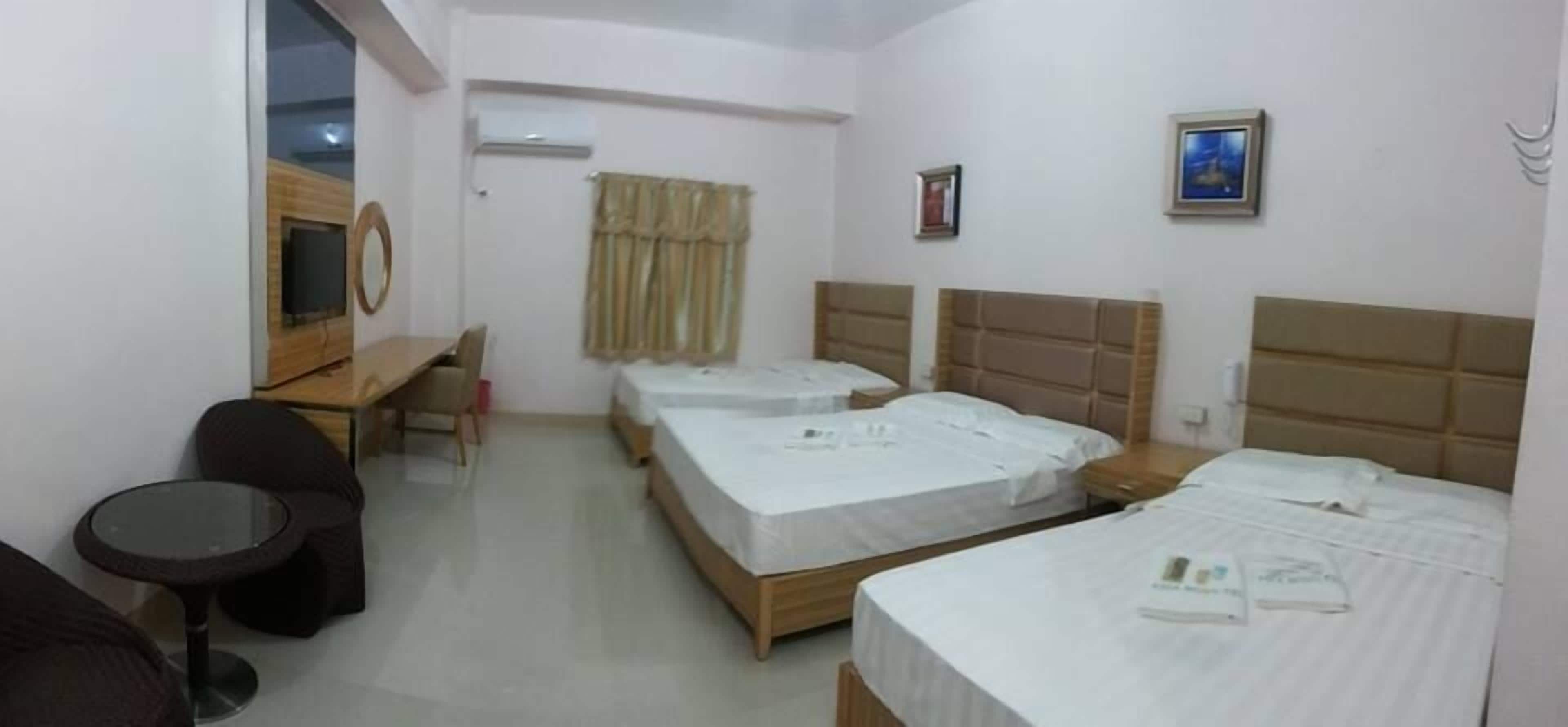 A hotel room with three single beds, a desk with a chair, a small round table, and a TV.