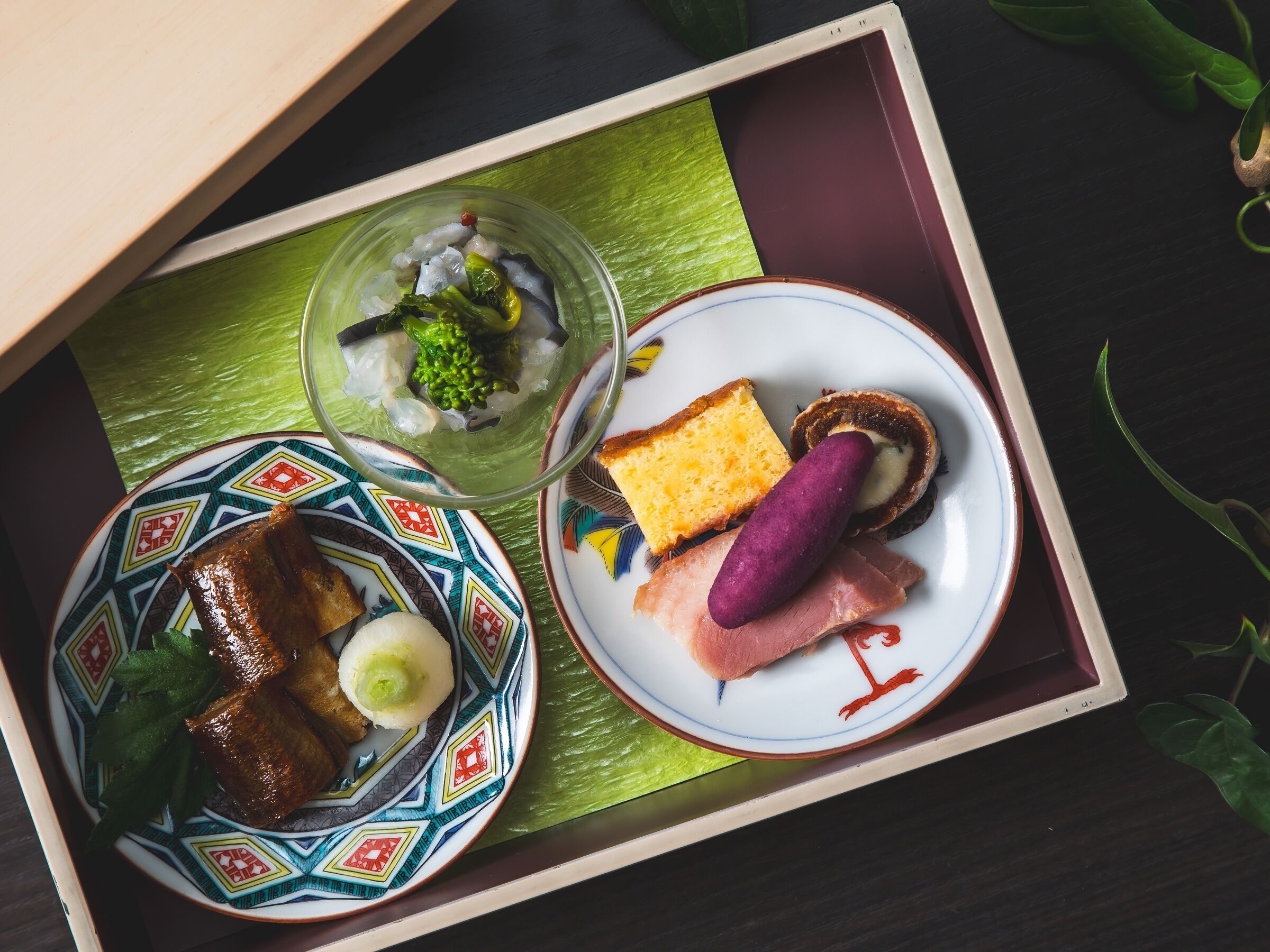 A wooden tray with a variety of dishes including a plate with a patterned design, a plate with a slice of bread, a slice of ham, and a purple sausage.
