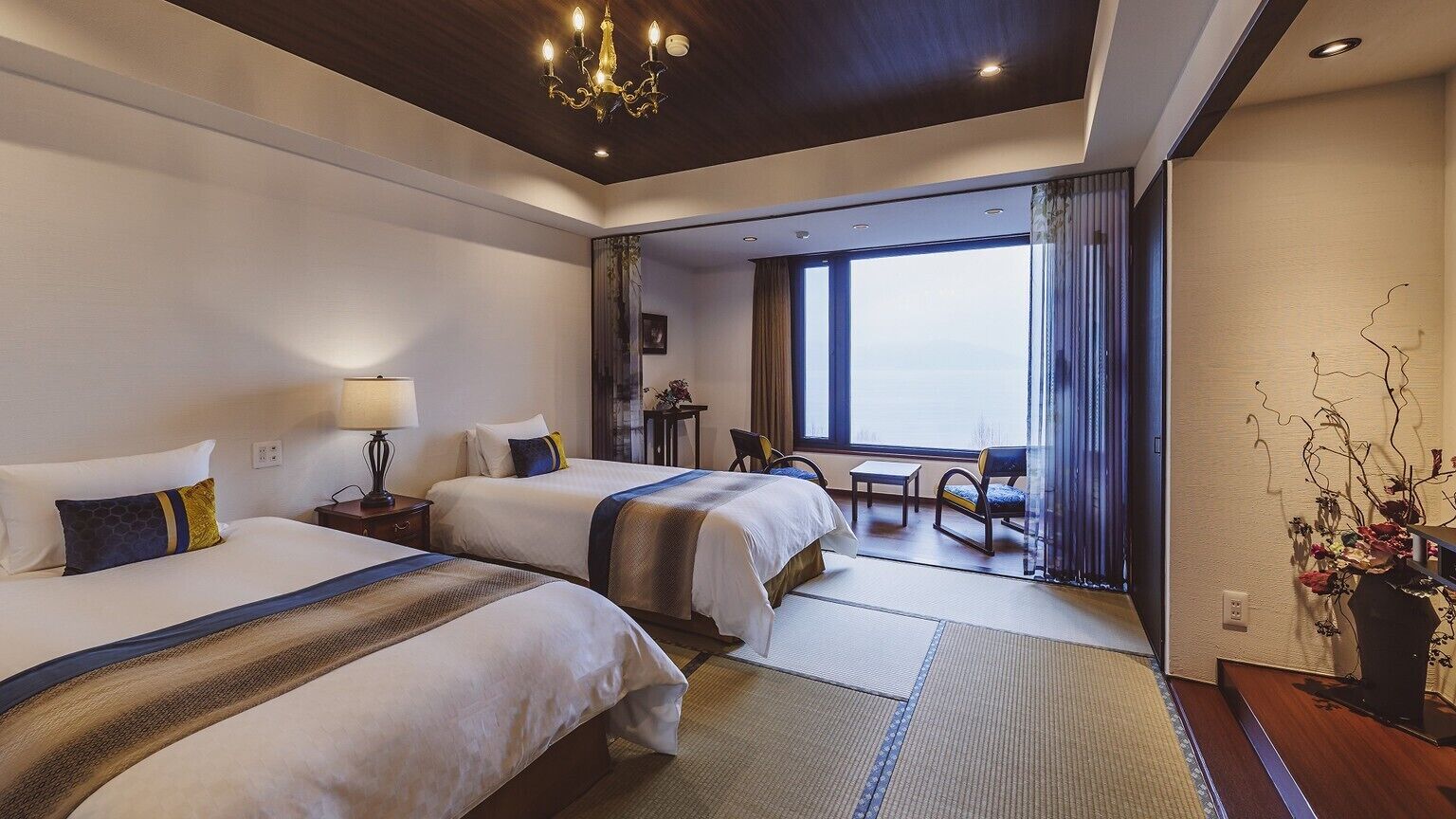 Une chambre d’hôtel avec deux lits, un lustre, une vue par une grande fenêtre et une petite table avec des chaises.