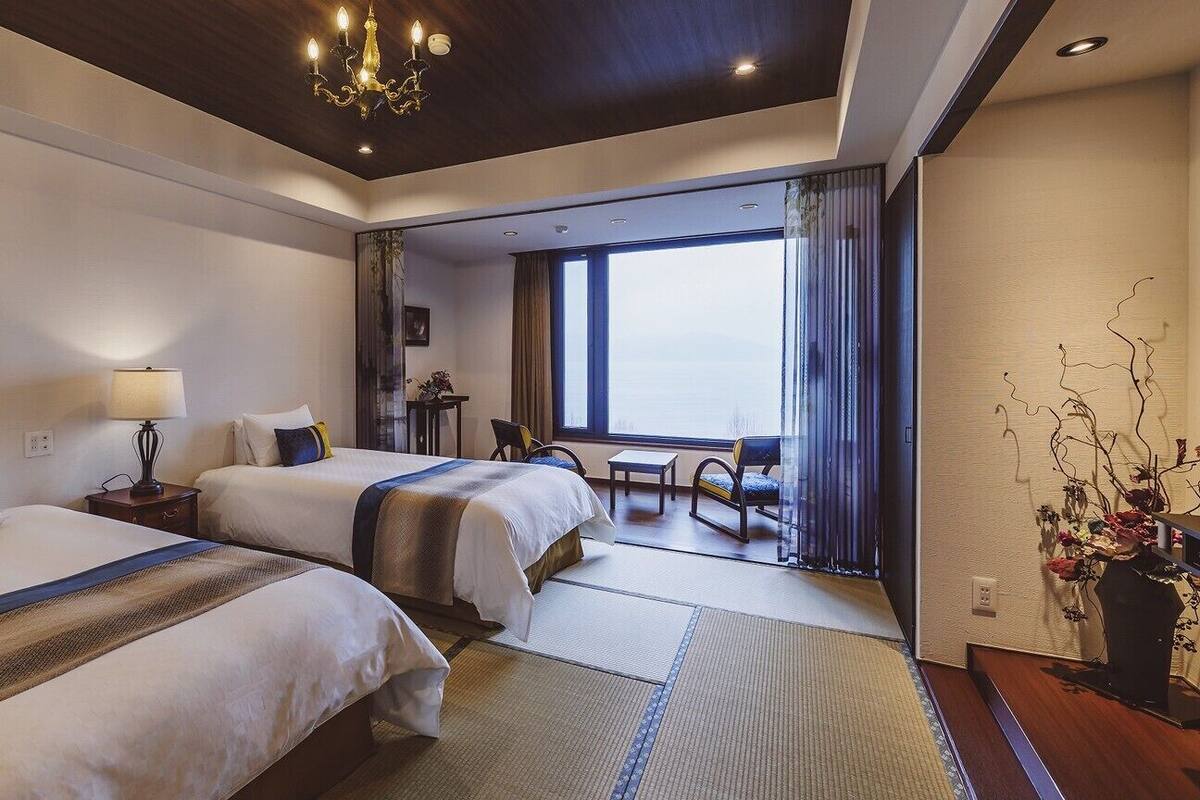 Une chambre d’hôtel avec deux lits, un lustre, une vue par une grande fenêtre et une petite table avec des chaises.