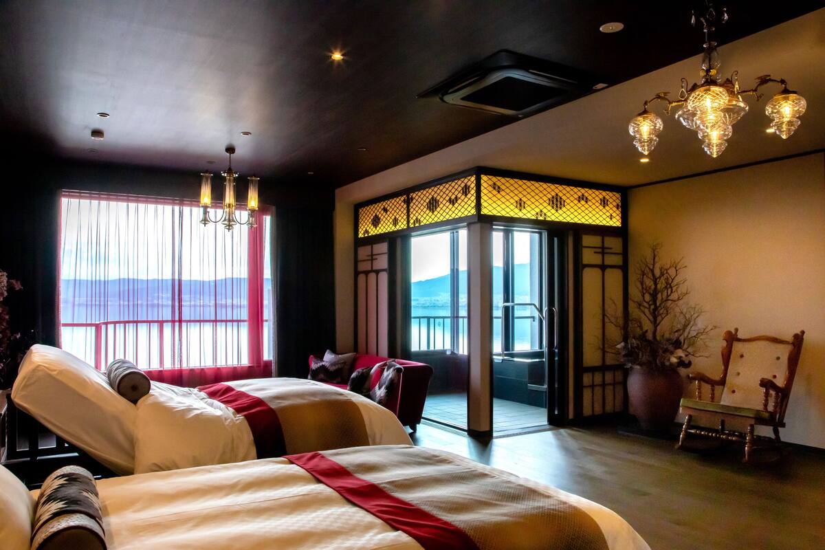 Une chambre d’hôtel avec deux lits, une grande fenêtre avec des rideaux, une vue sur les montagnes, un paravent japonais traditionnel et une chaise en bois.