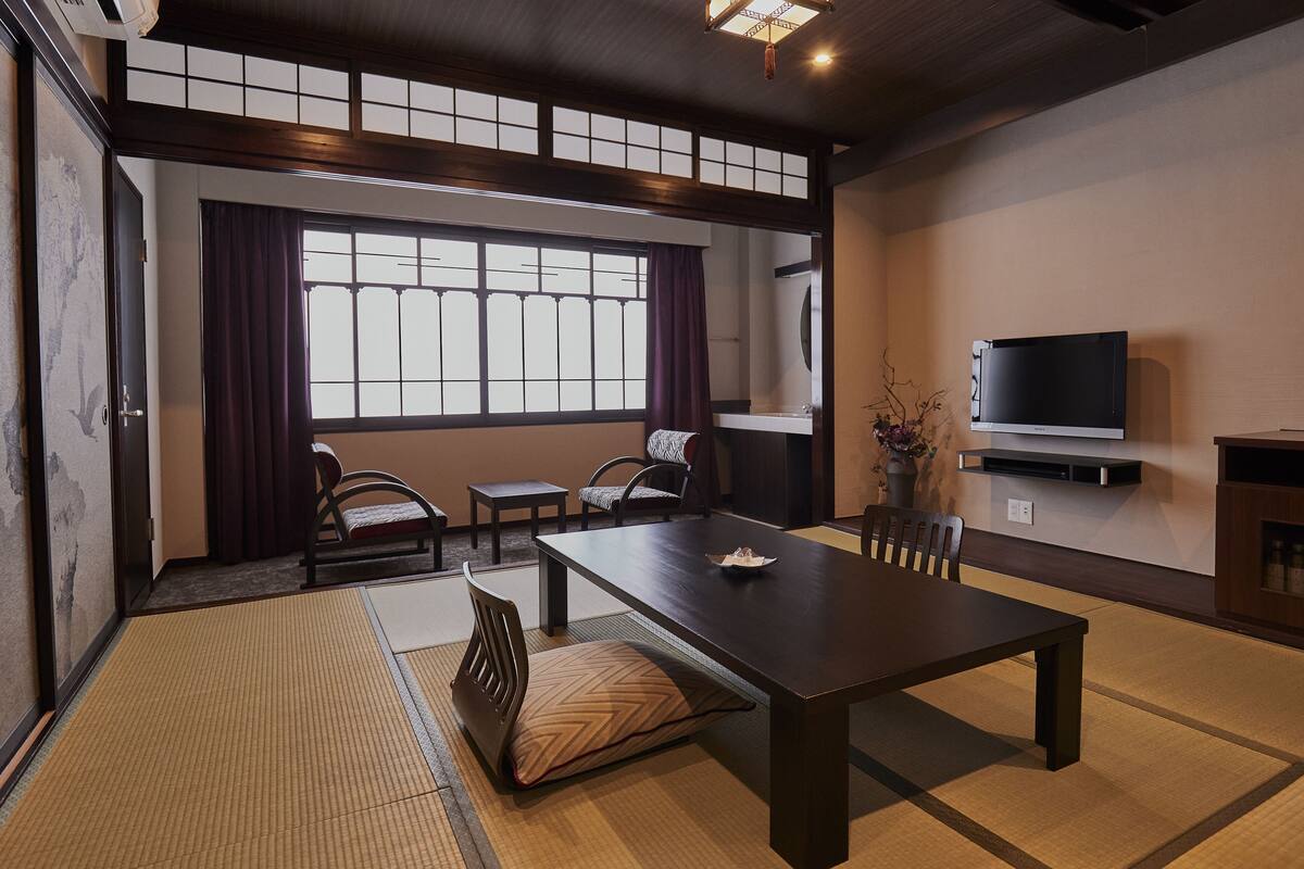 Une pièce de style japonais traditionnel, avec un sol recouvert de tatamis, une table basse, deux chaises, un téléviseur et une fenêtre avec des rideaux.