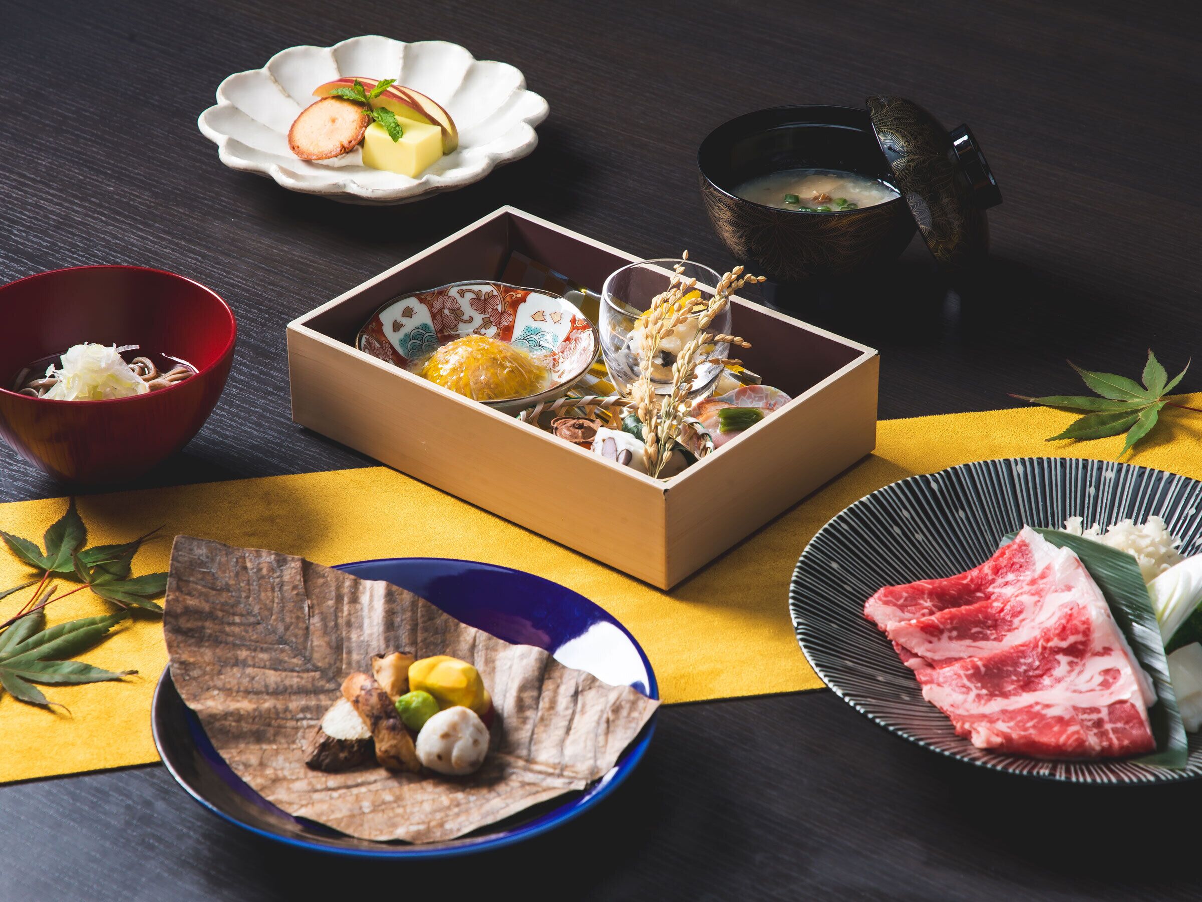 A traditional Japanese meal setup with a wooden box containing various foods, a bowl of soup, and plates with meat and vegetables.