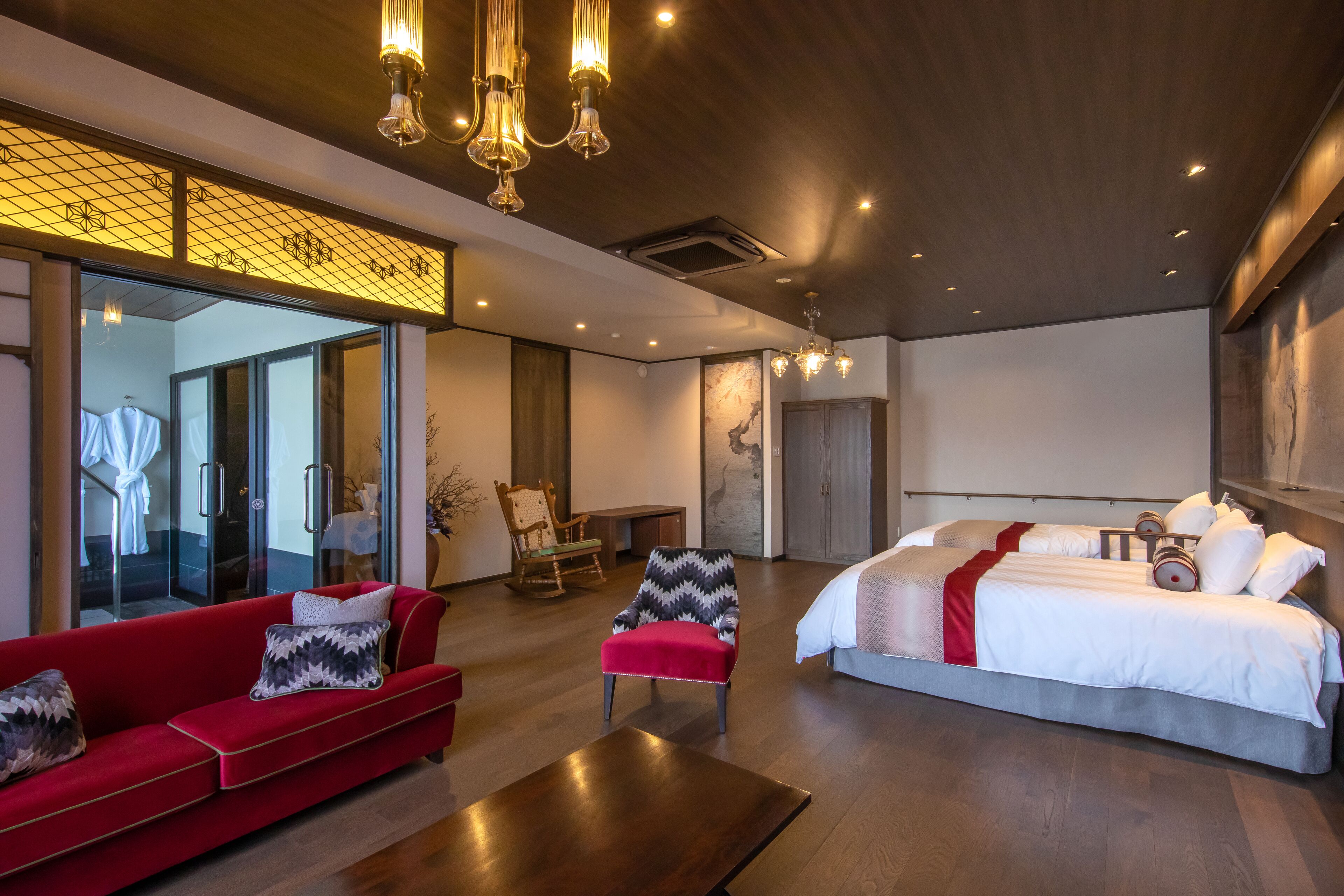A modern hotel room with a large bed, a red sofa, a wooden coffee table, and a seating area with a chair and a small table.