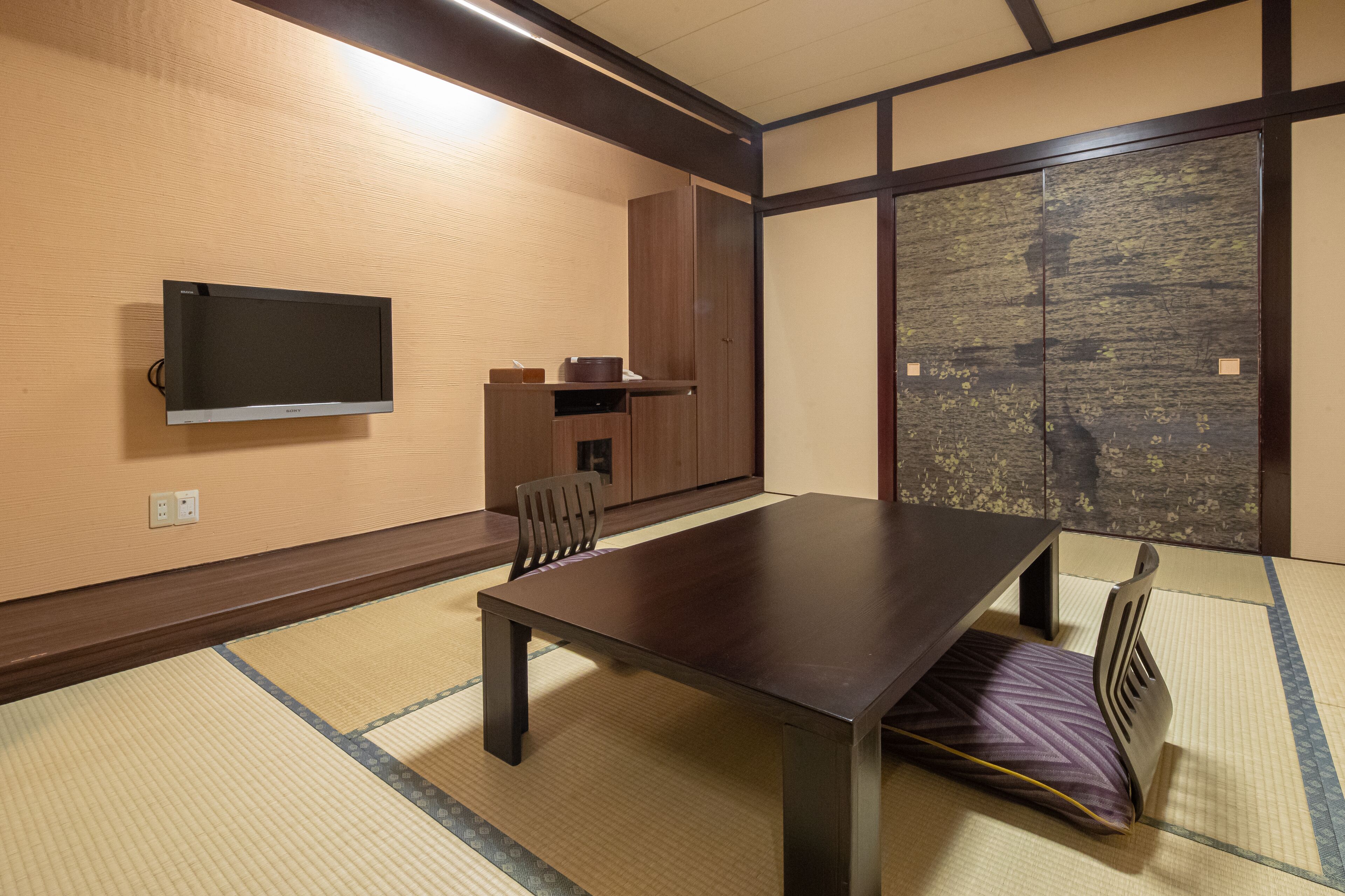 A traditional Japanese room with tatami flooring, a low table, chairs, a television, and a sliding door with a view.