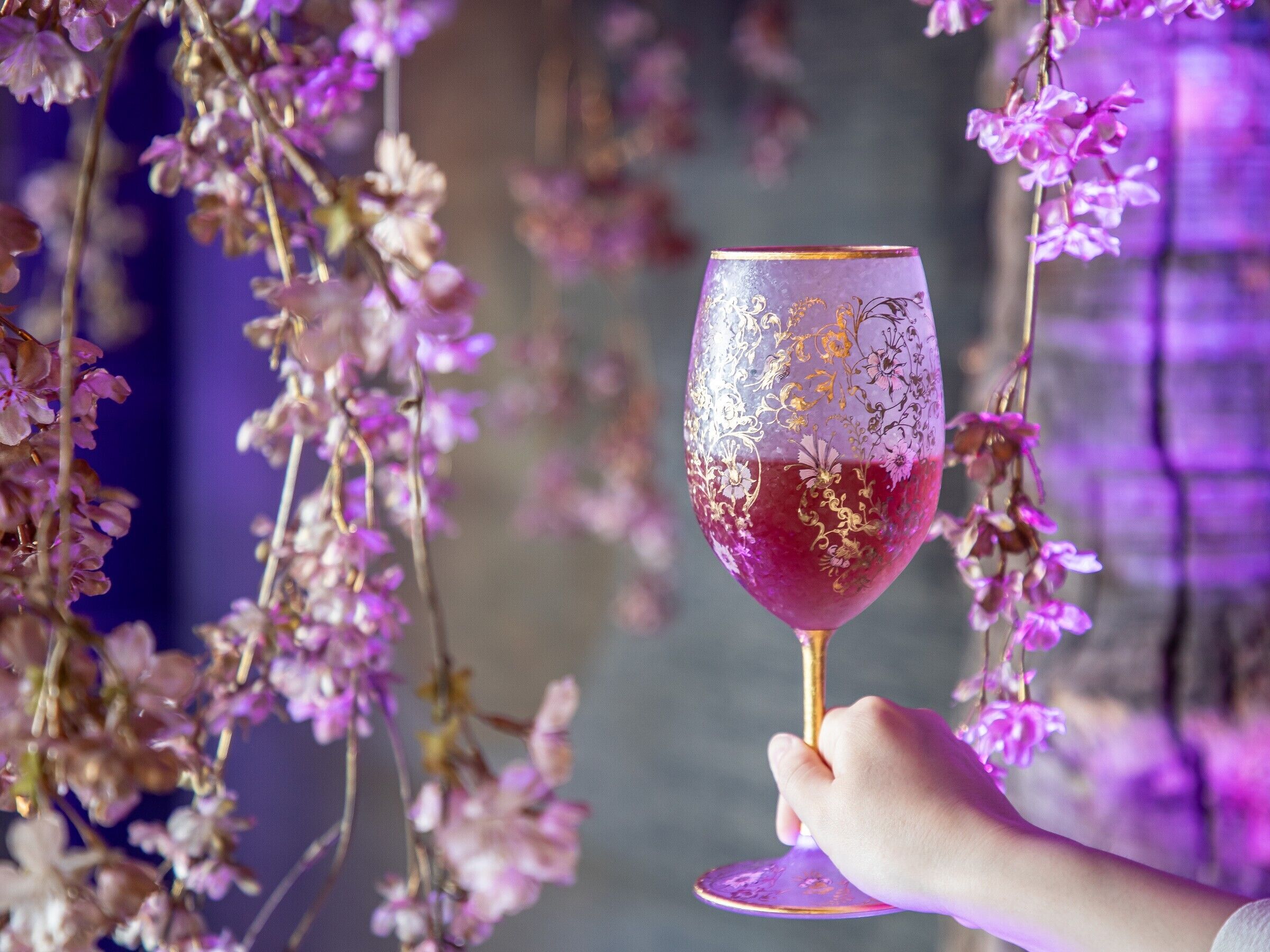 A hand holding a wine glass with a gold pattern, surrounded by purple flowers.
