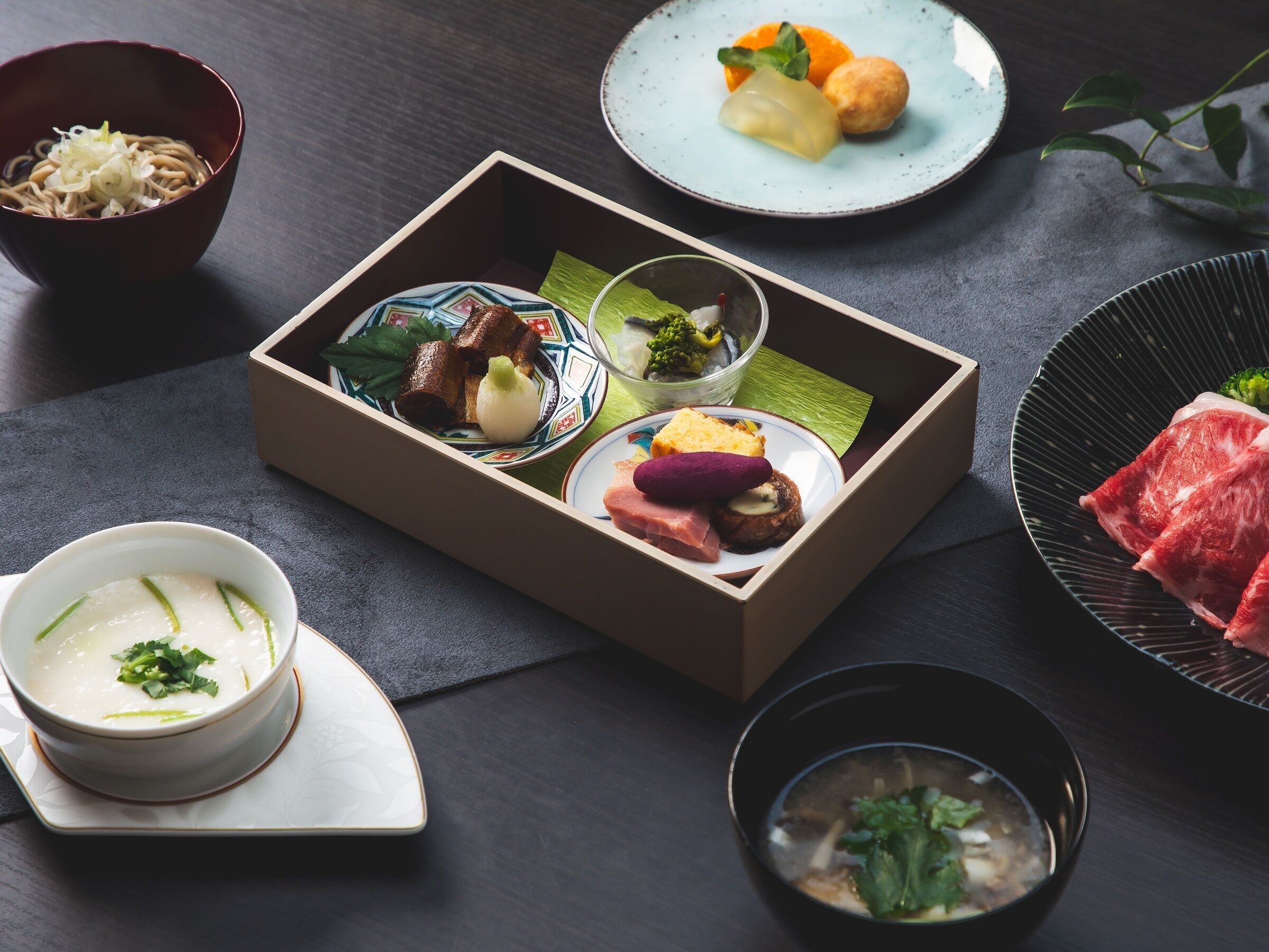 Un repas japonais traditionnel, composé de différents plats, notamment un bol de soupe, une assiette de viande et une boîte contenant des légumes marinés, des légumes frais et un petit plat de tofu.