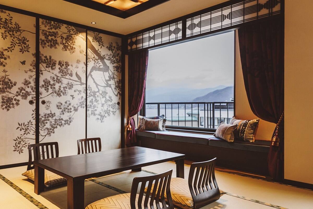Une pièce de style traditionnel, avec un sol recouvert de tatamis, une table basse et des chaises, un paravent shoji orné de motifs floraux, et une vue sur la mer par une grande fenêtre.