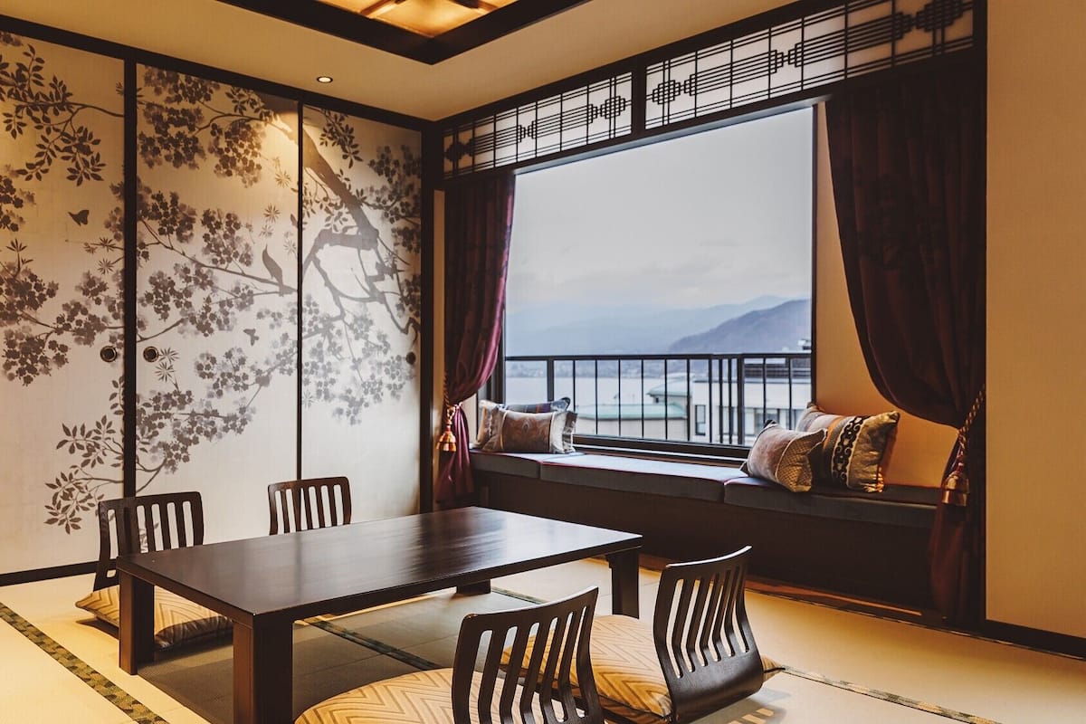 Une pièce de style traditionnel, avec un sol recouvert de tatamis, une table basse et des chaises, un paravent shoji orné de motifs floraux, et une vue sur la mer par une grande fenêtre.