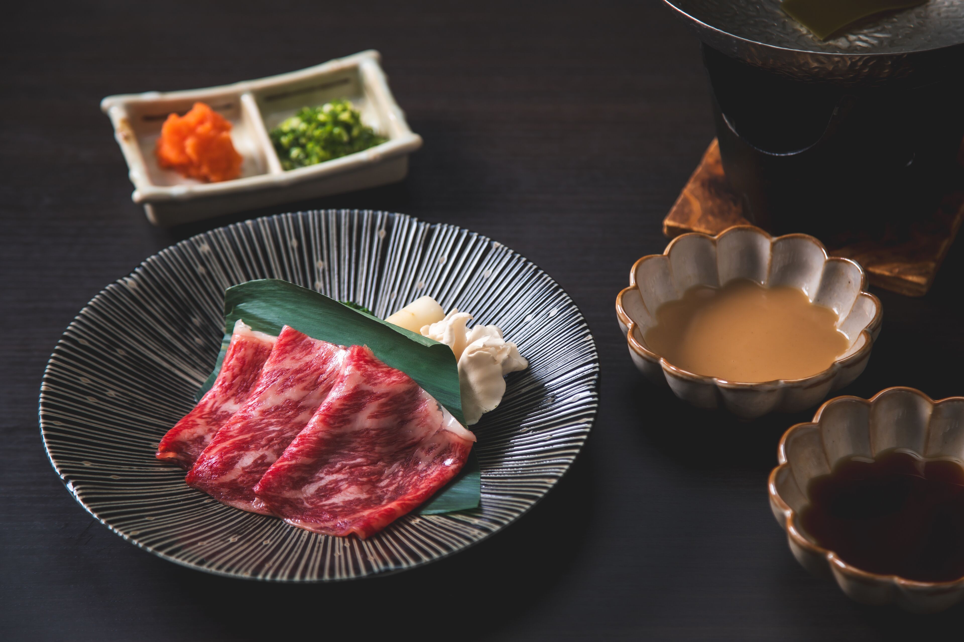 A plate with slices of meat, a bowl of soup, and a cup of tea on a dark table.