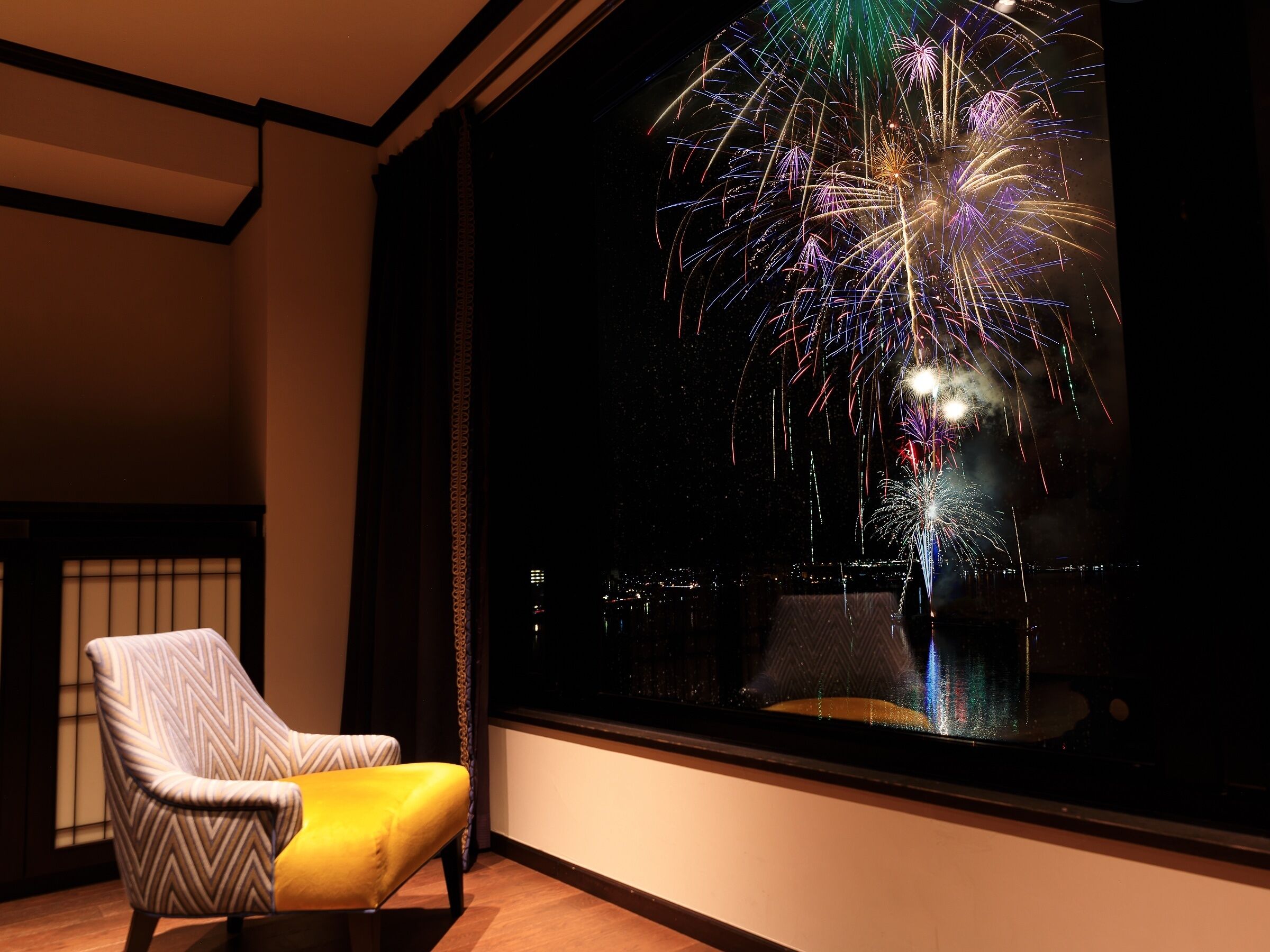 A room with a large window overlooking a cityscape, a chair, and a view of fireworks.