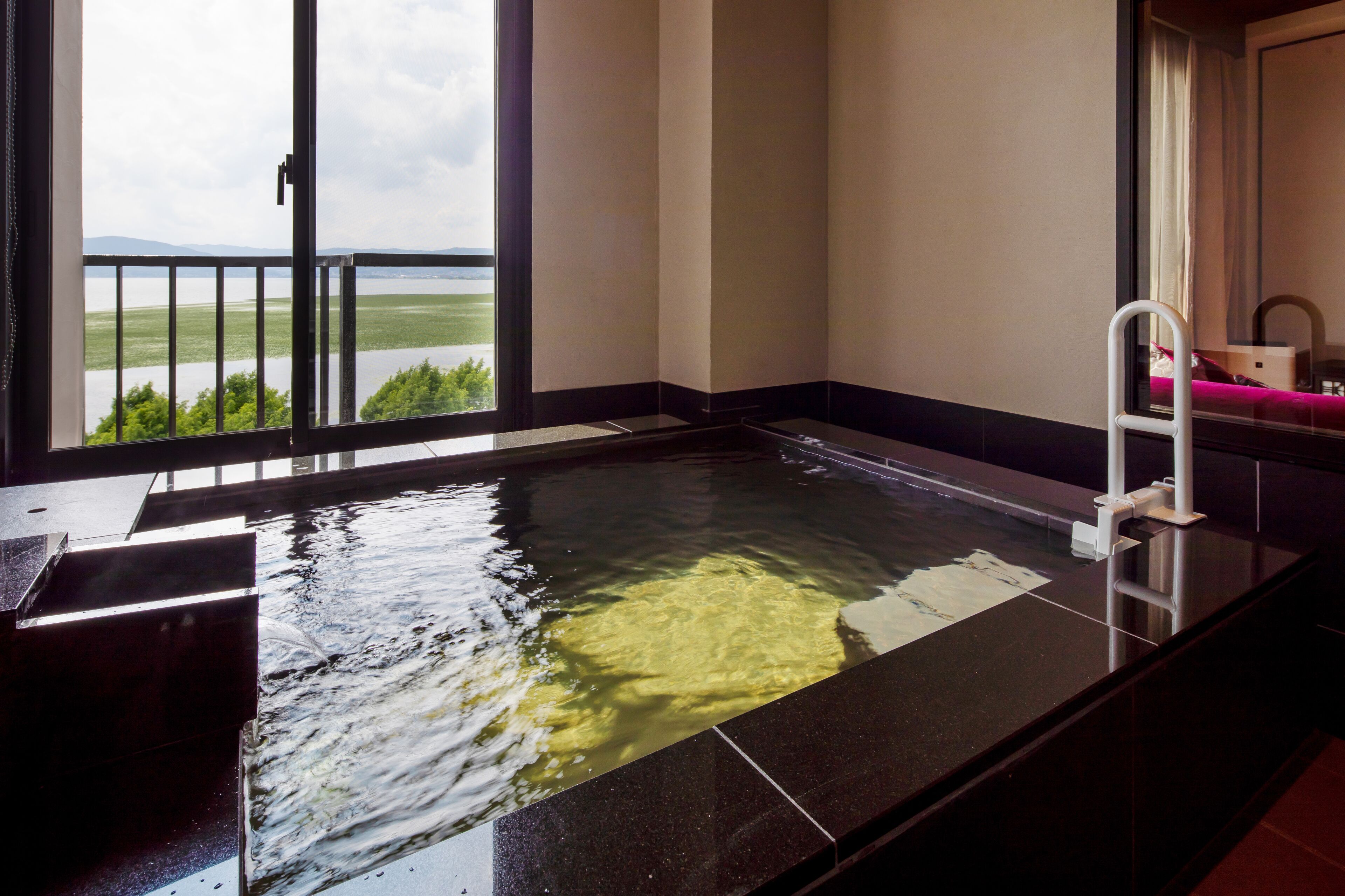 A room with a black-tiled hot tub, a white ladder, and a view of a lake through a large window.