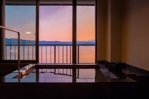 A room with a large window overlooking a body of water and distant mountains during sunset.