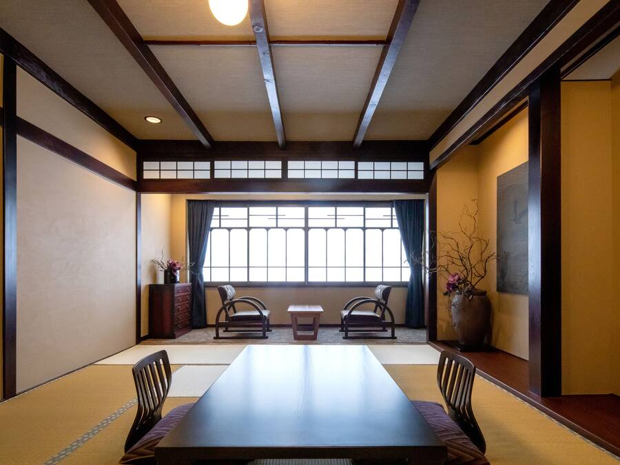 Una habitación tradicional japonesa con suelo de tatami, una mesa baja, dos sillas y un ventanal con cortinas.