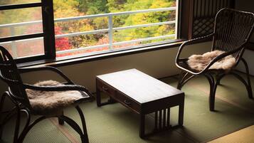 A traditional Japanese room with tatami flooring, a low wooden table, two dark wooden chairs with fur cushions, and a window with a view of autumn foliage.