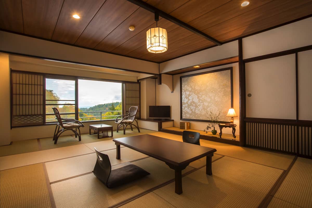 Ein traditioneller japanischer Raum mit Tatami-Boden, einem niedrigen Tisch, Stühlen, einem Fernseher und einem großen Fenster mit Blick ins Freie.