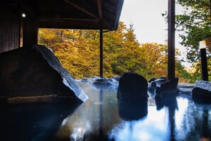 Een traditionele Japanse onsen met houten constructie, een natuurlijke rotsvijver en herfstbladeren op de achtergrond.