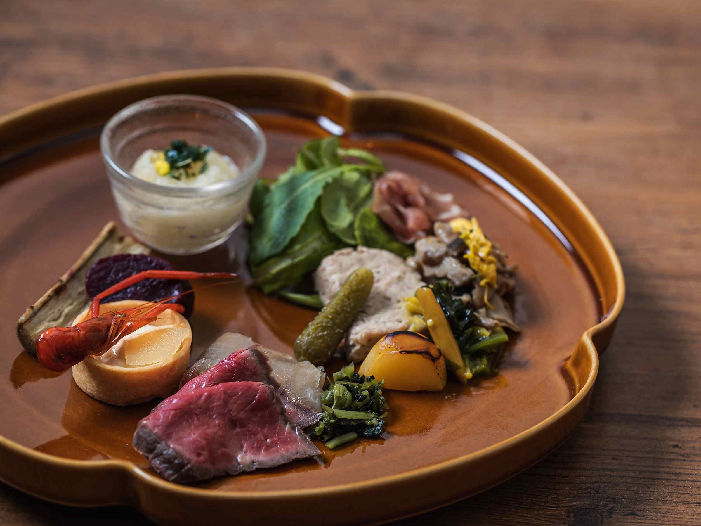 Un plato con diversos alimentos, incluyendo carne, verduras y un recipiente pequeño con salsa.