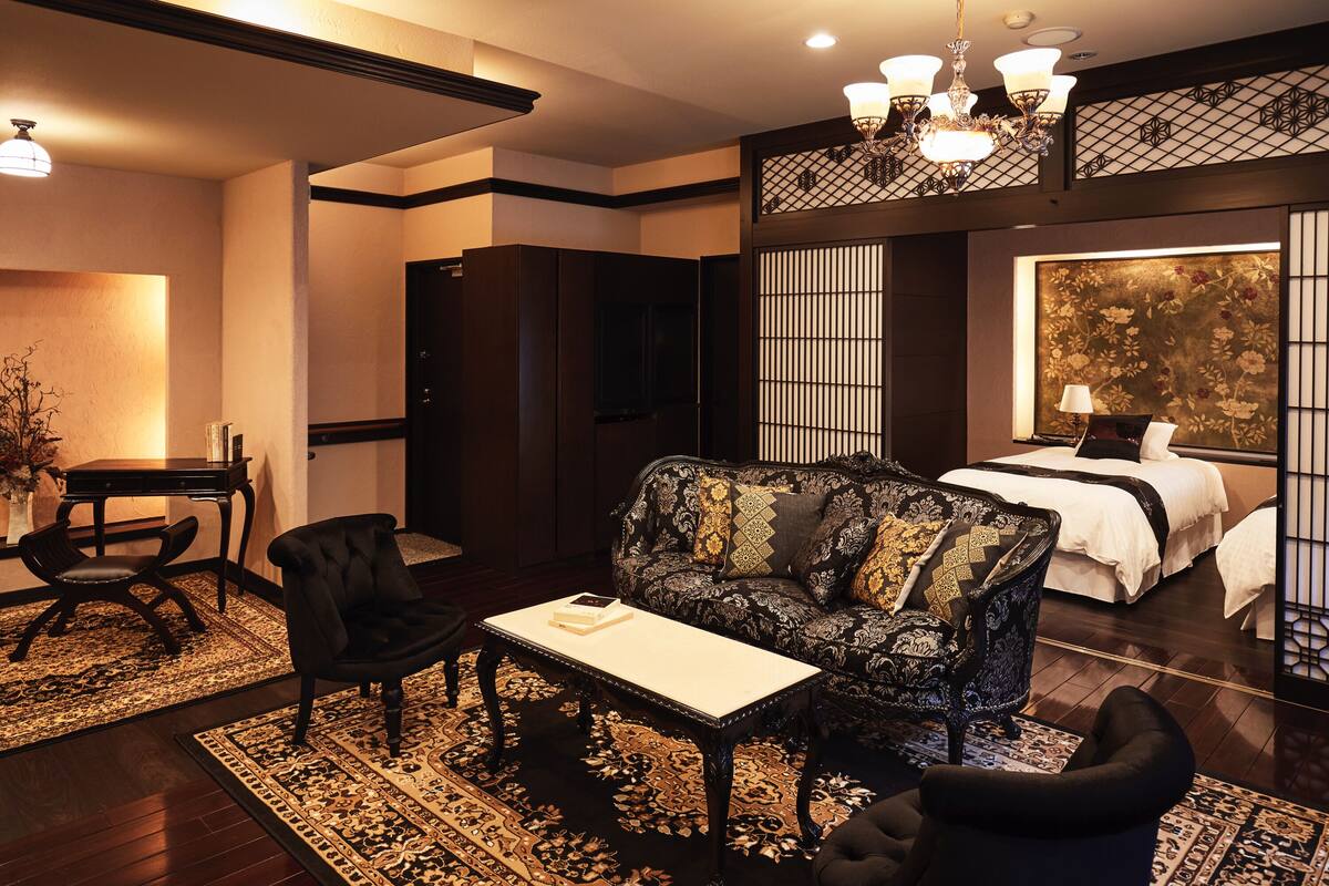 Ein Hotelzimmer mit traditioneller japanischer Einrichtung, inklusive Tatami-Boden, einem niedrigen Tisch mit einem Buch, einer Couch und einem Bett mit großem Fenster.