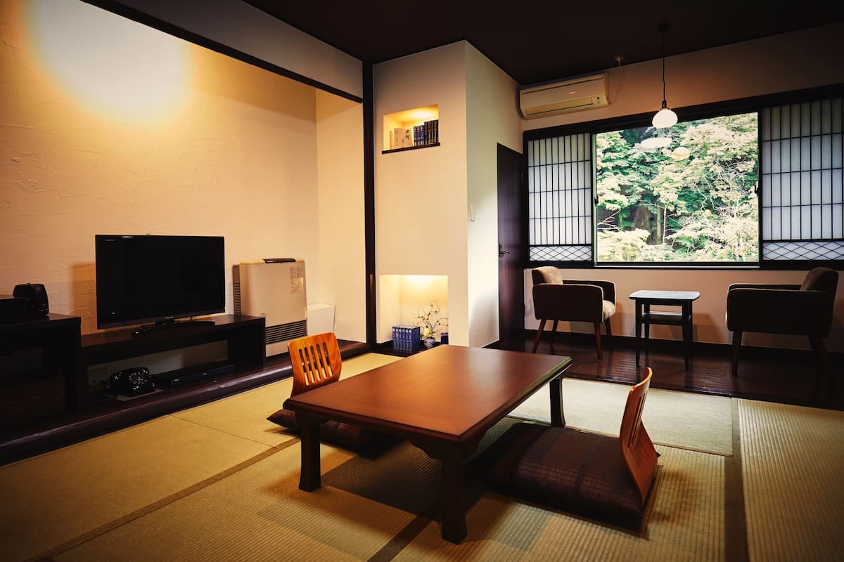 Ein traditioneller japanischer Raum mit Tatami-Boden, einem niedrigen Tisch, Stühlen, einem Fernseher und einem großen Fenster mit Blick auf Bäume.