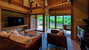 A spacious living room with a large TV, comfortable seating, and a view of a lush garden through large windows.