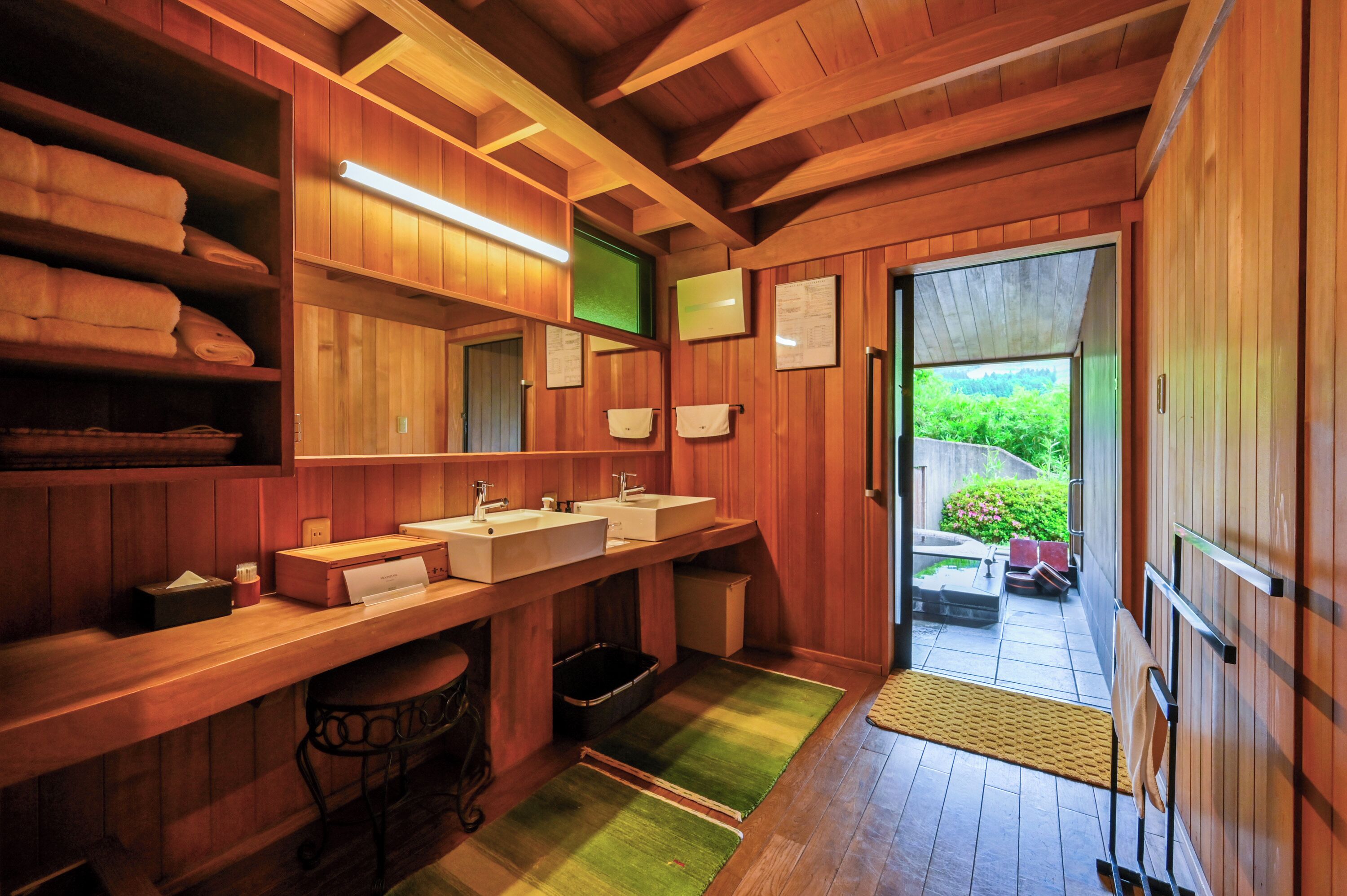 Une salle de bain en bois avec deux lavabos, des étagères avec des serviettes et une vue sur la verdure à l’extérieur.