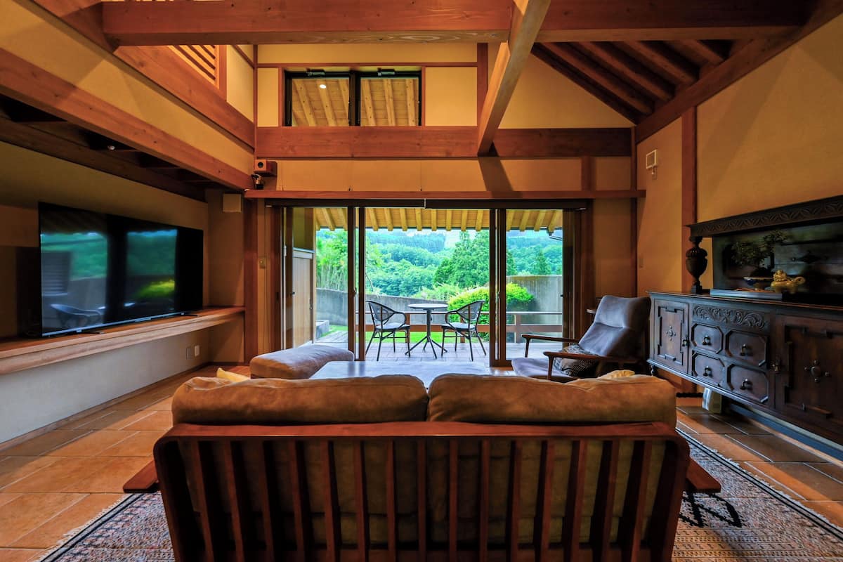Un salon équipé d’un téléviseur, de meubles en bois, d’un coin repas avec des chaises et offrant une vue sur la verdure à l’extérieur.