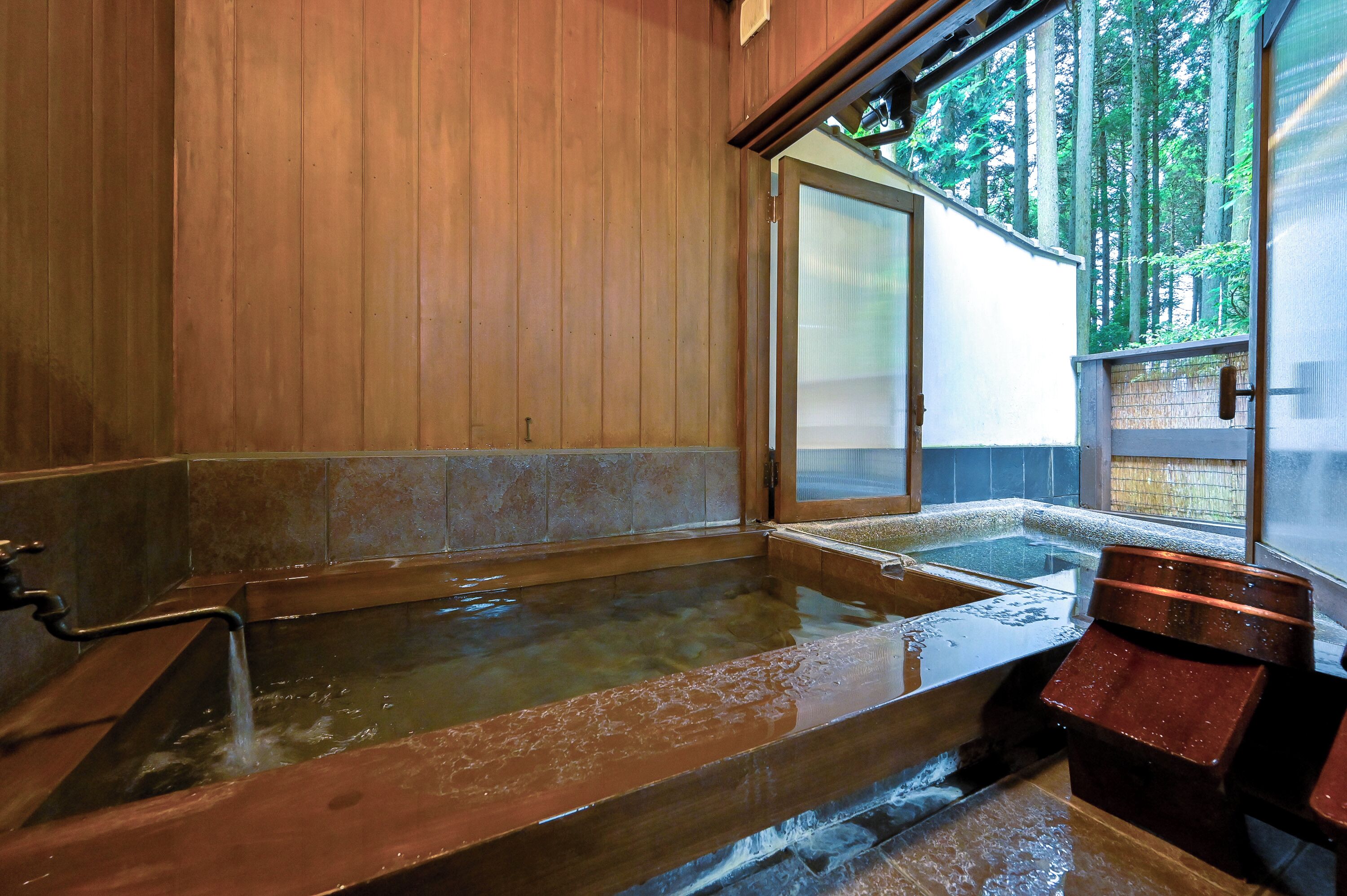 A traditional wooden bathhouse with multiple hot tubs and a sliding glass door leading to a forested area.