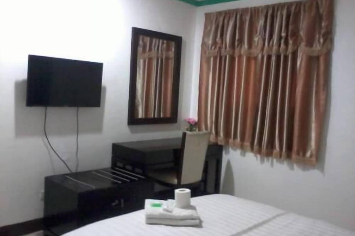 Habitación de hotel con cama, escritorio, silla, televisor y ventana con cortinas.