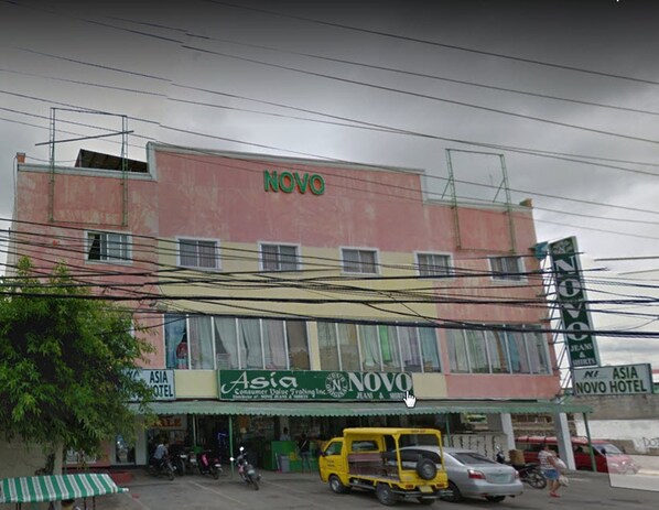 Un hotel con un letrero destacado que dice 'NOVO', un local con rótulos que indican 'Asia Consumer Value Trading Inc' y 'ASIA HOTEL', y un auto-rickshaw amarillo adelante.