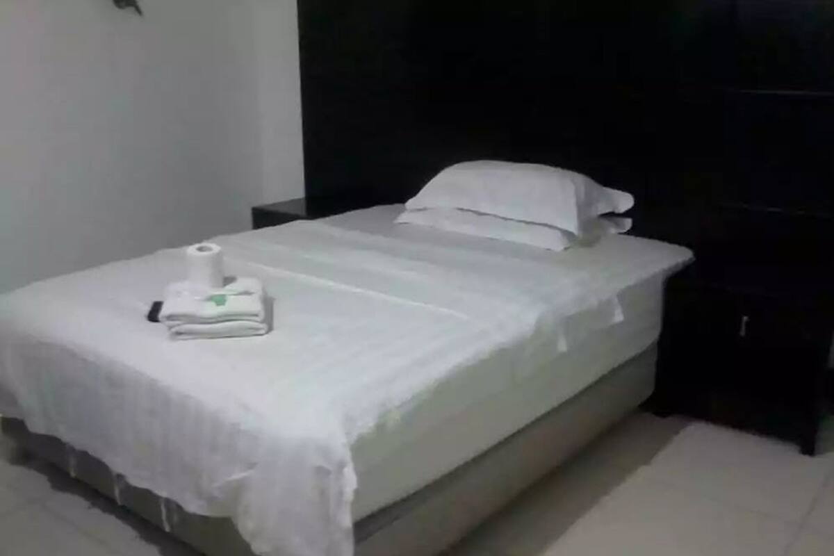 Una cama individual con una colcha blanca y almohadas, una toalla doblada y una mesita de noche con una lámpara.