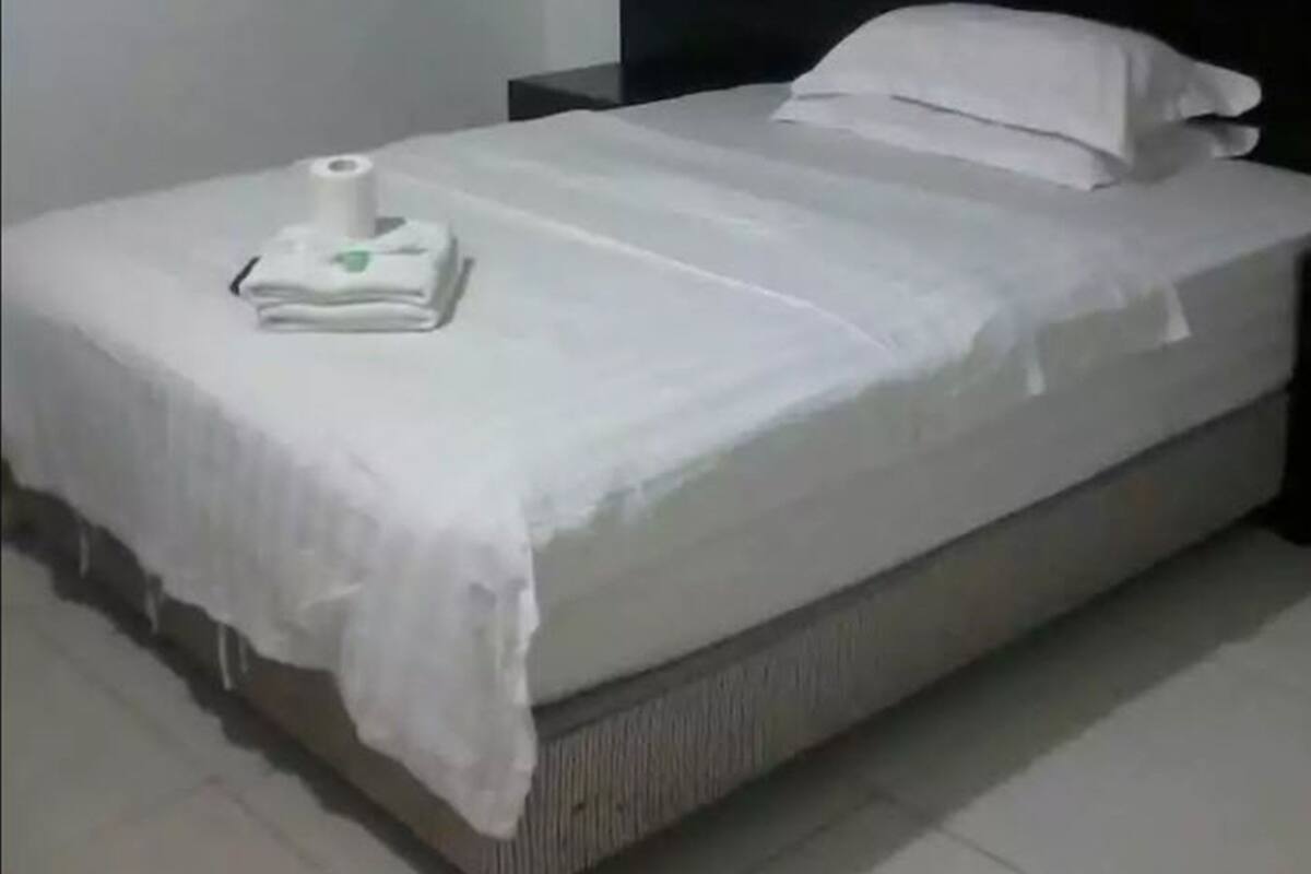 Una cama bien tendida con edredón blanco y almohadas, una mesita de noche con una lámpara y un aire acondicionado de pared.