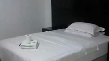 Una cama bien tendida con edredón blanco y almohadas, una mesita de noche con una lámpara y un aire acondicionado de pared.