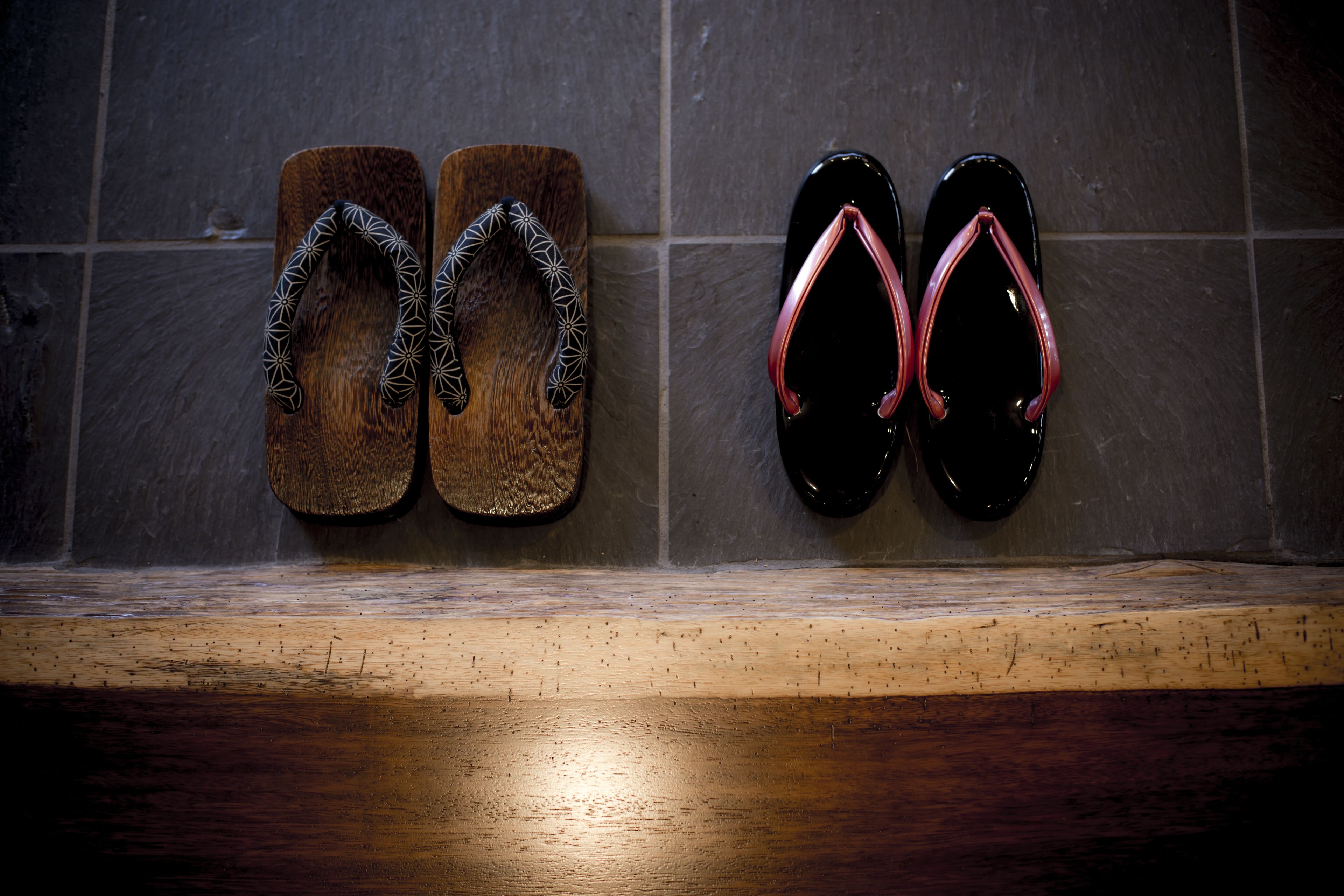A pair of wooden slippers and a pair of black flip-flops hanging on a tiled wall.
