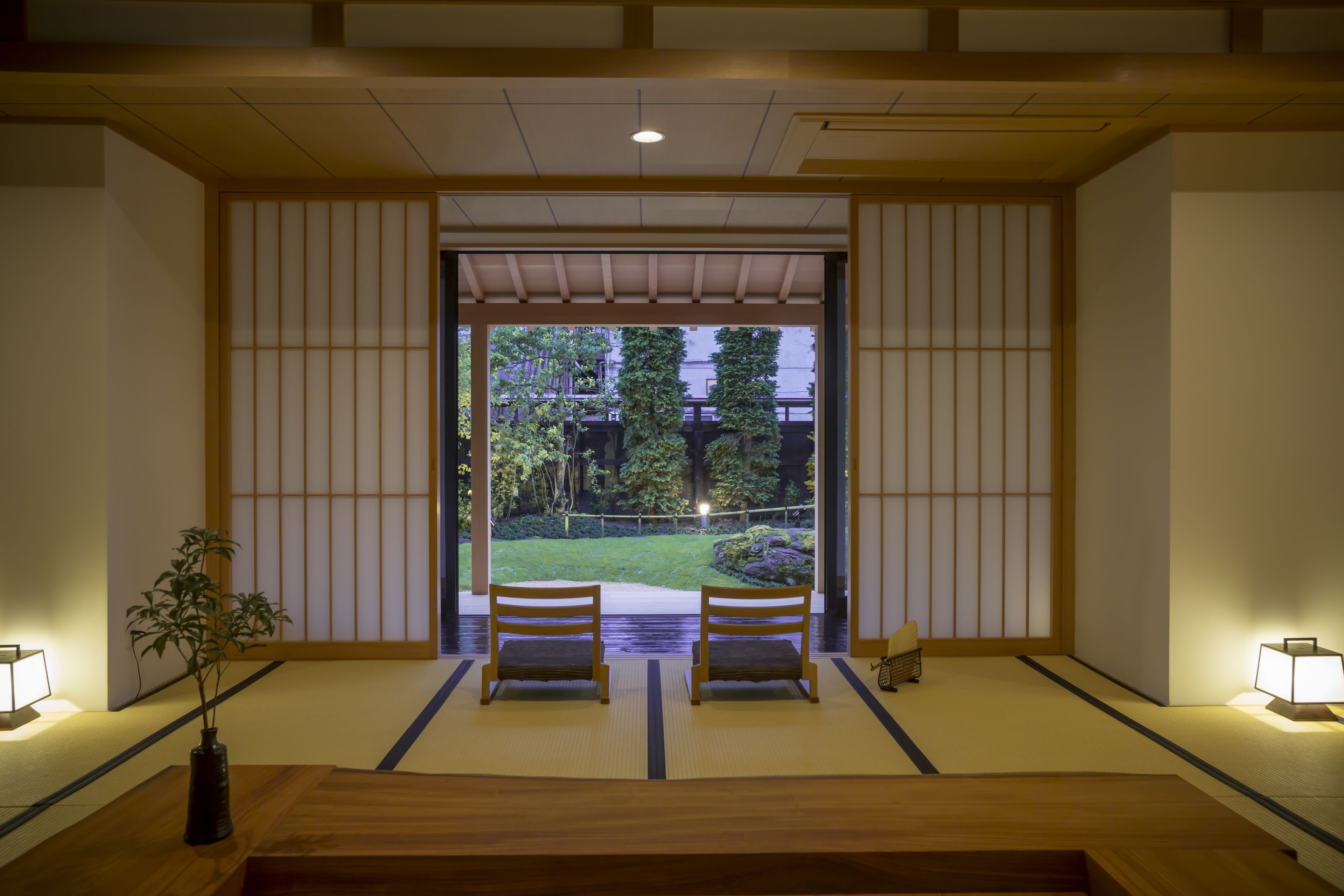 A traditional Japanese room with tatami flooring, a low table, two chairs, and a view of a garden through a shoji screen.
