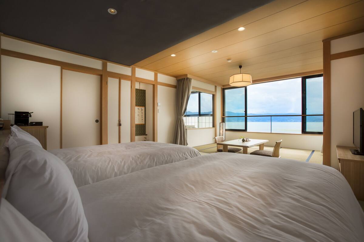 A hotel room with two beds, a small table, a chair, a TV, and a large window with a view of the sea.