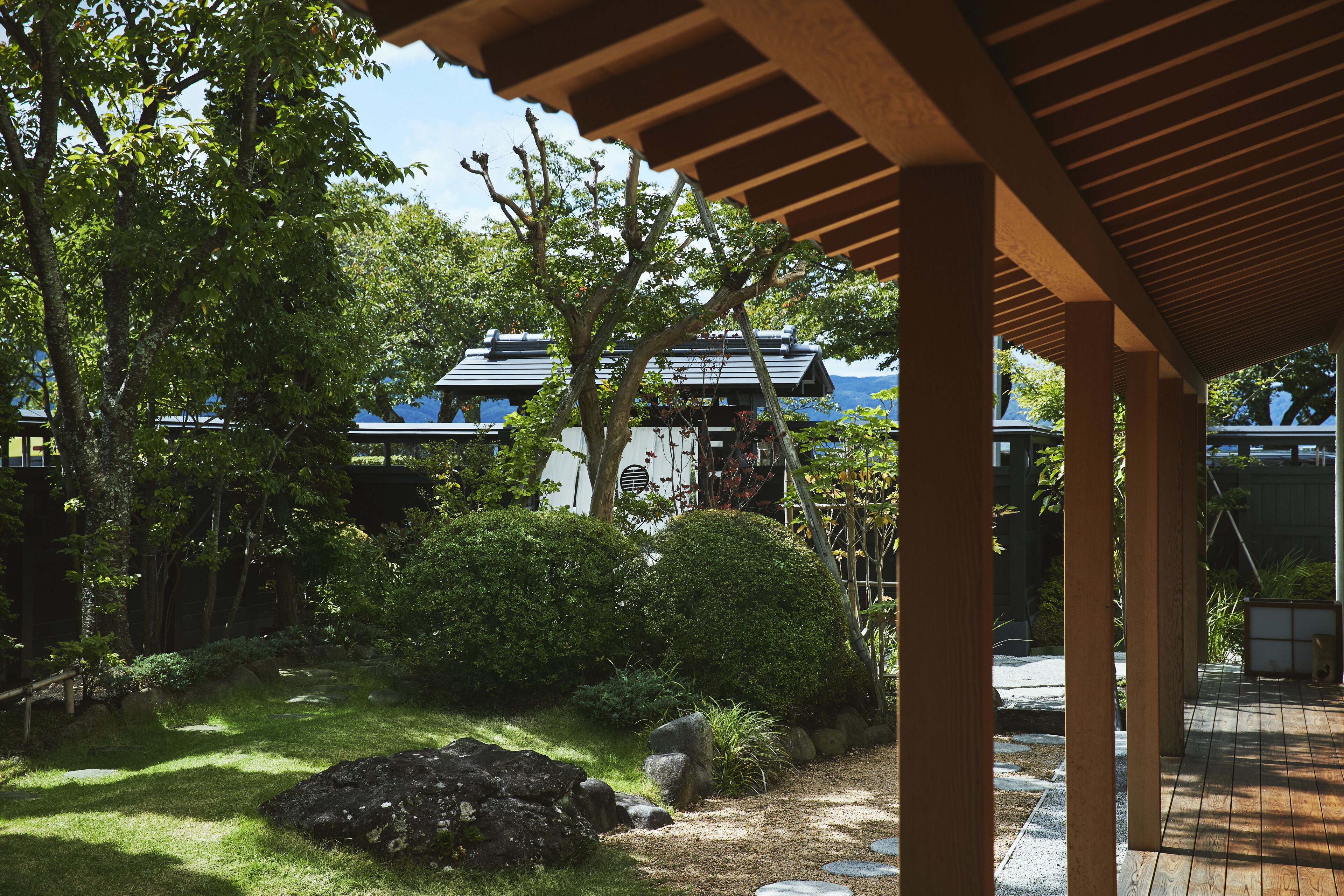 A traditional Japanese garden with a wooden walkway, rocks, and lush greenery.