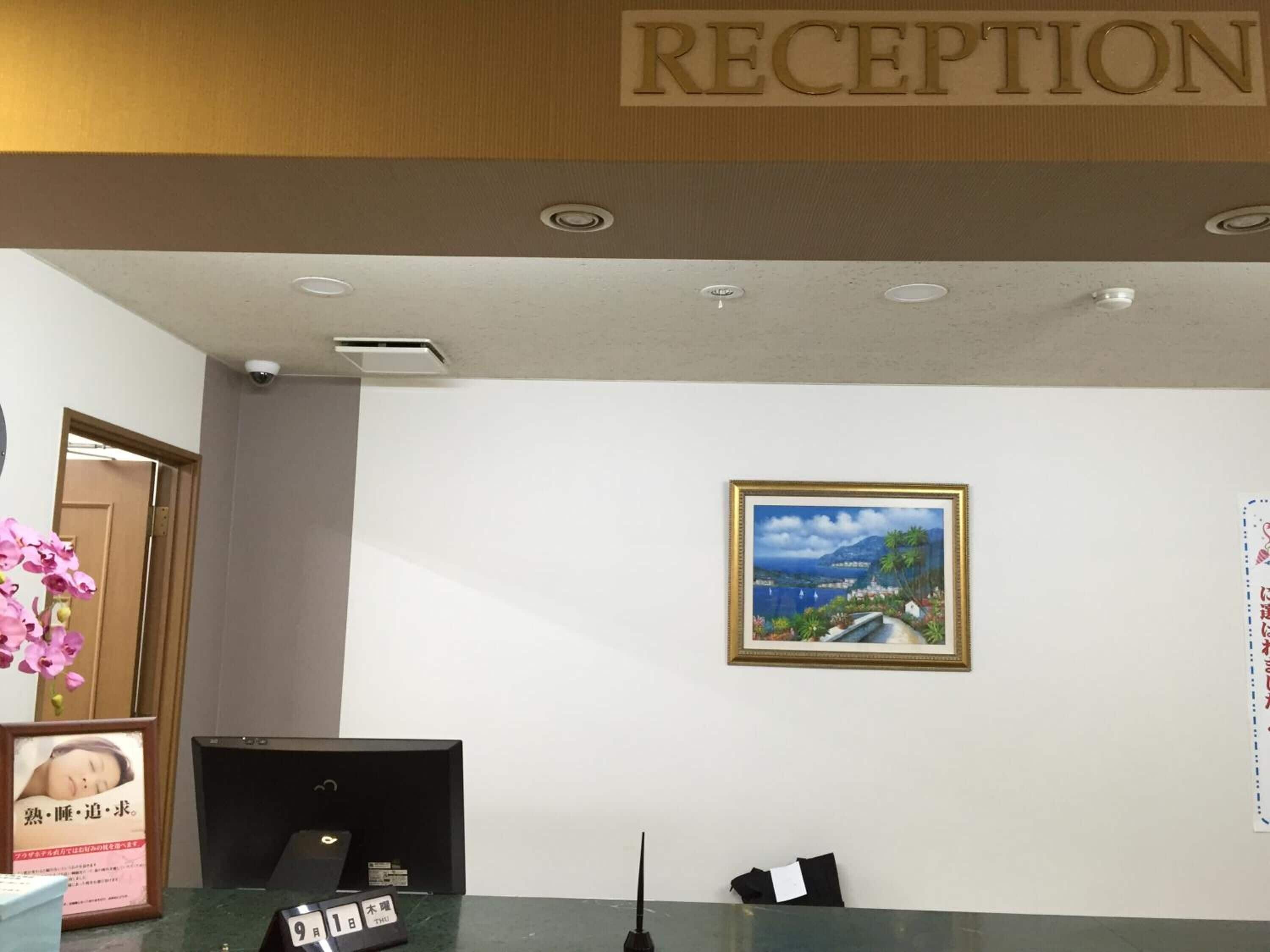 Un'area reception con un cartello sopra che indica ‘RECEPTION’.  Una cornice ospita un dipinto raffigurante un paesaggio con acqua e vegetazione. Si vede una scrivania nera con un monitor computerizzato, e un piccolo cartello con caratteri giapponesi è posizionato sulla scrivania.