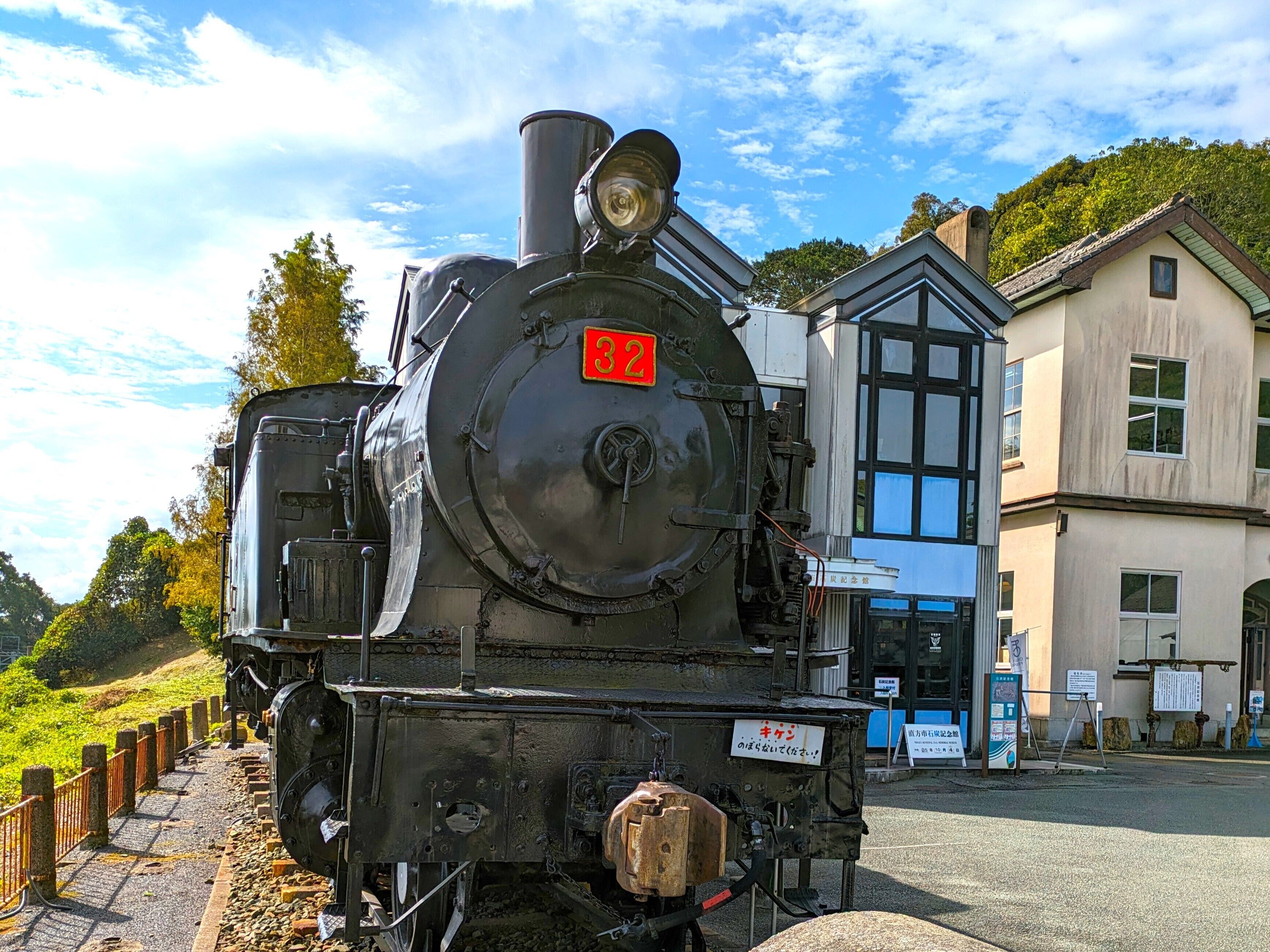 Una locomotora de vapor con el número 32, estacionada junto a un edificio con ventanas grandes y un cartel.
