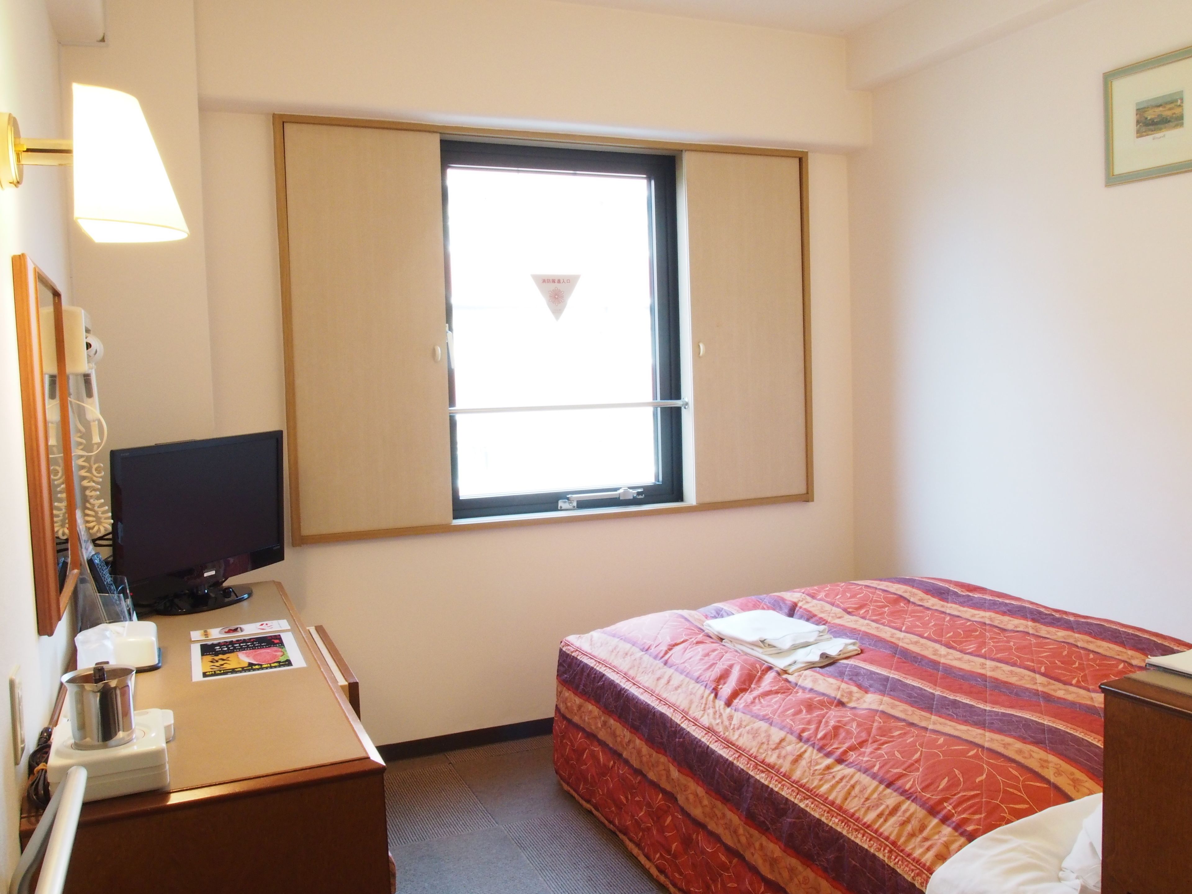 A hotel room with a bed, a desk with a computer, a window, and a wall-mounted TV.