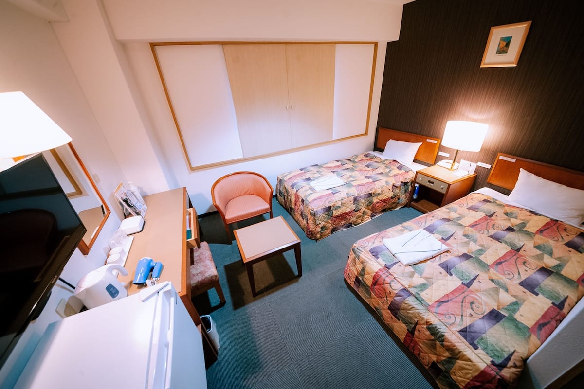A hotel room with two beds, a desk, a chair, a small table, and a wardrobe.
