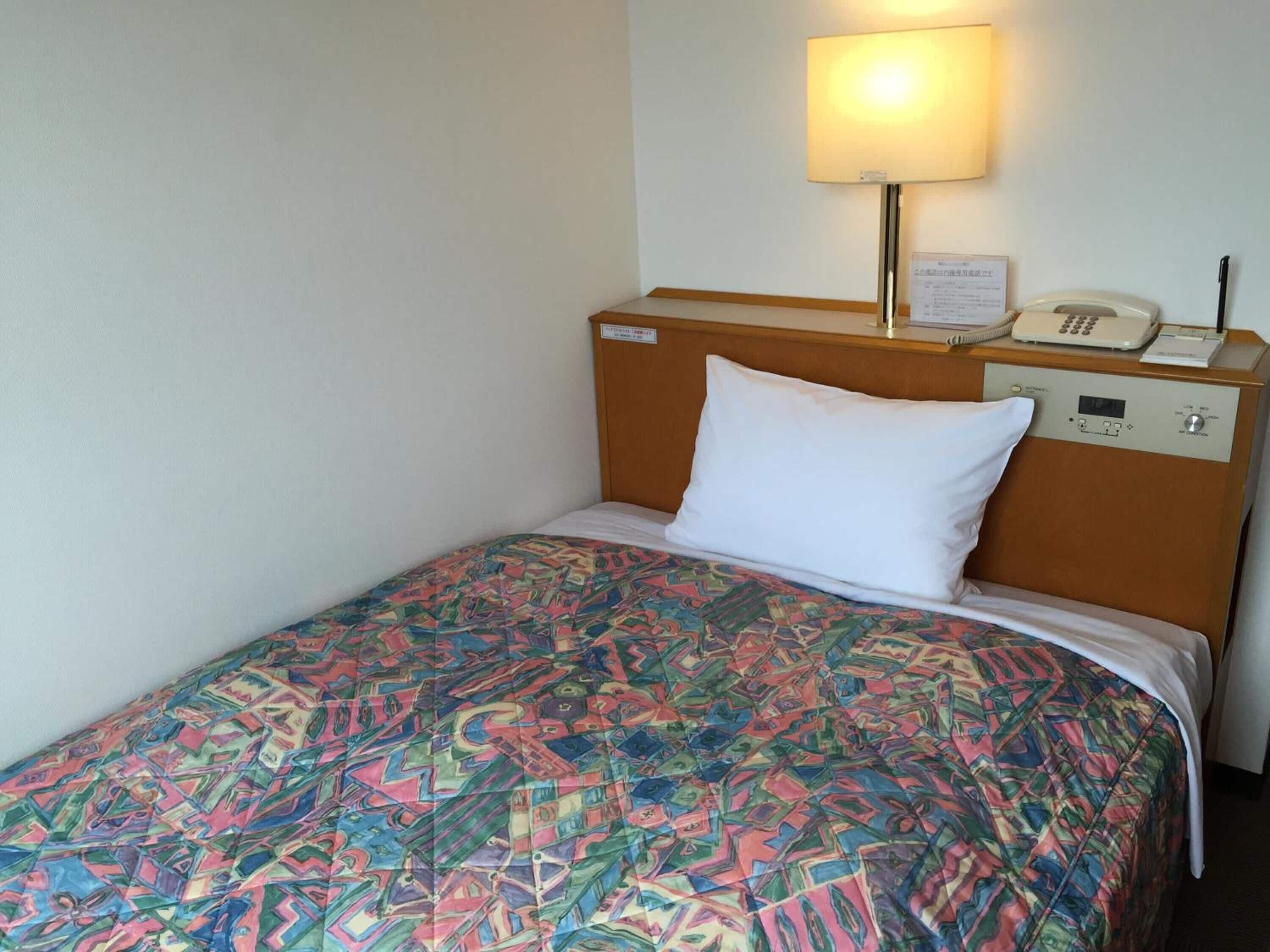 Una habitación de hotel con cama, lámpara de noche, teléfono y panel de control en la cabecera.
