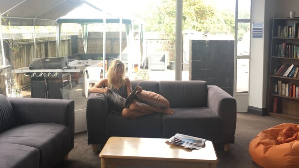 A living room with a sofa, a coffee table, and a bean bag. There is a bookshelf with books and a window with a view of the outside. - BoBs Hostel (Taupo)