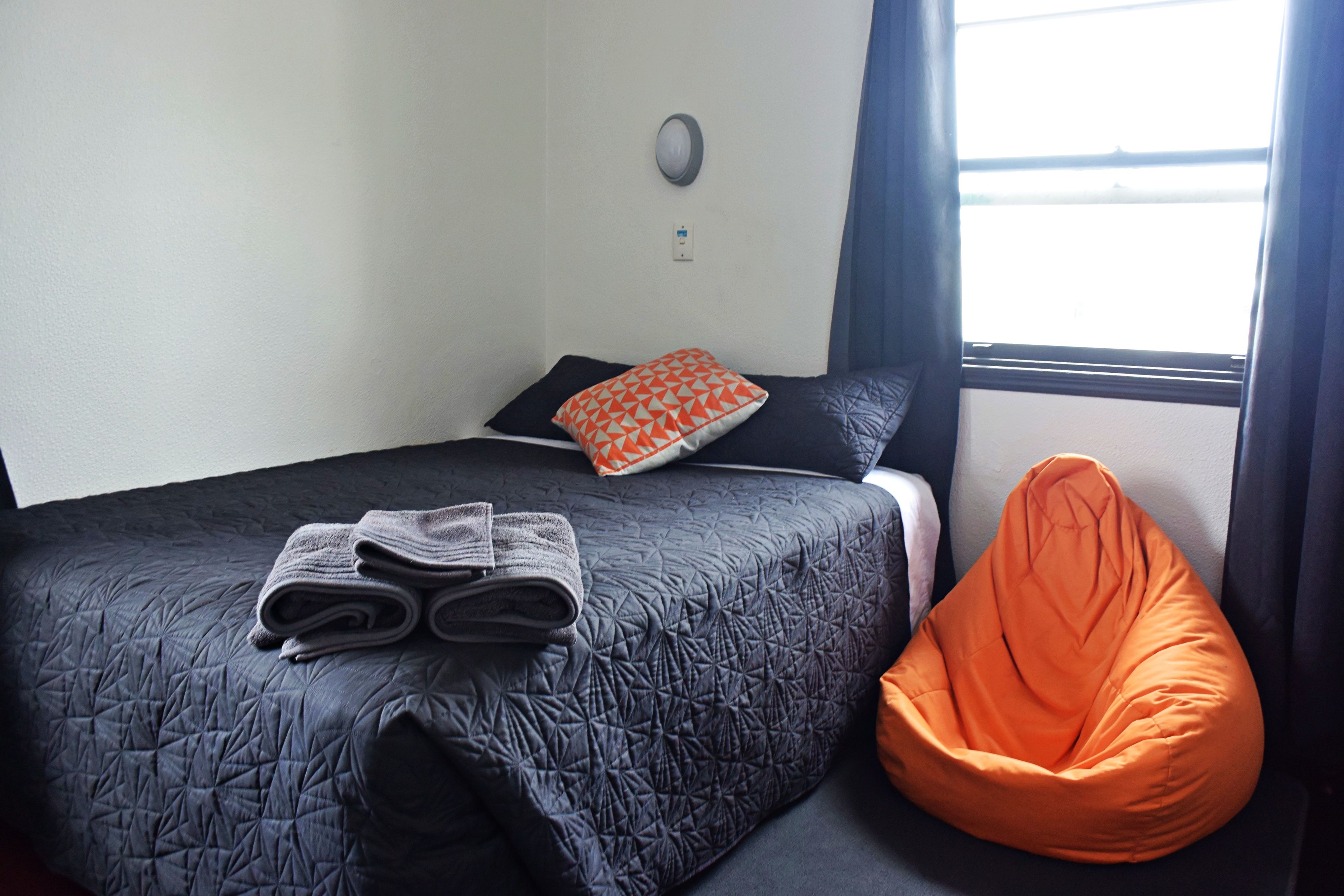 A bed with a dark quilt, an orange bean bag chair, and a pillow with a geometric pattern.