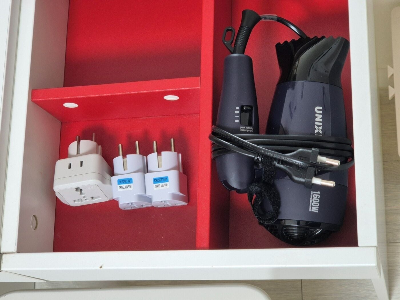 A drawer with a red interior containing a travel adaptor, two plugs, and a black device labeled "UNIX" with a cable.